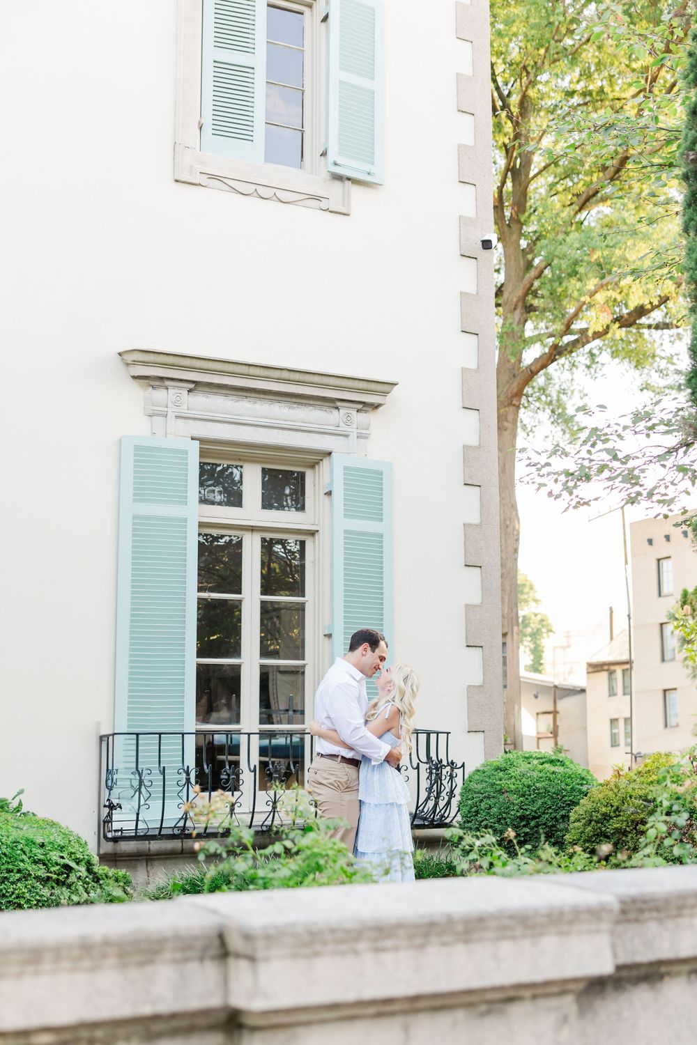 A couple embraces on a balcony with light blue shutters and classic architecture in the background.