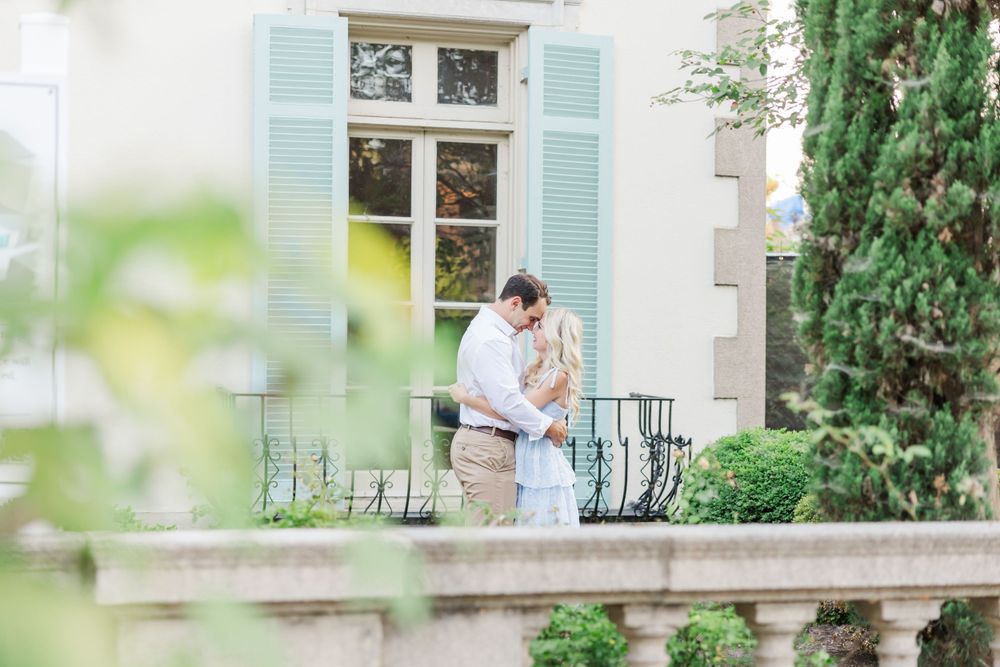 Romantic moment captured through greenery on an elegant balcony with pale blue shutters.