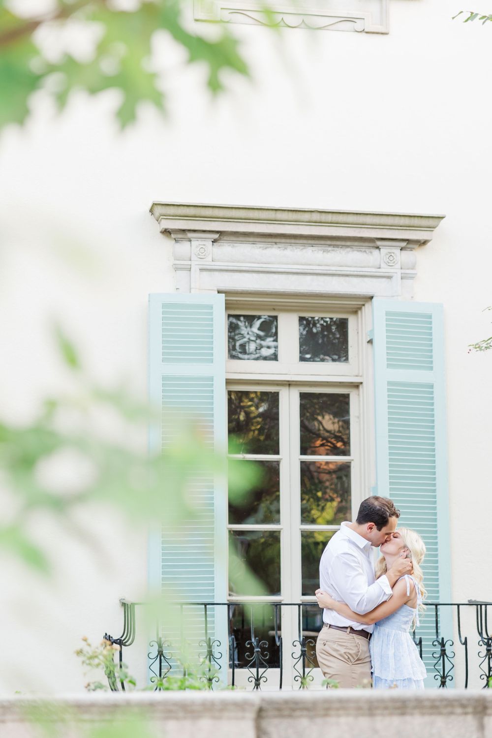 Tender embrace outside a French-style window with pale blue shutters and ornate balcony.