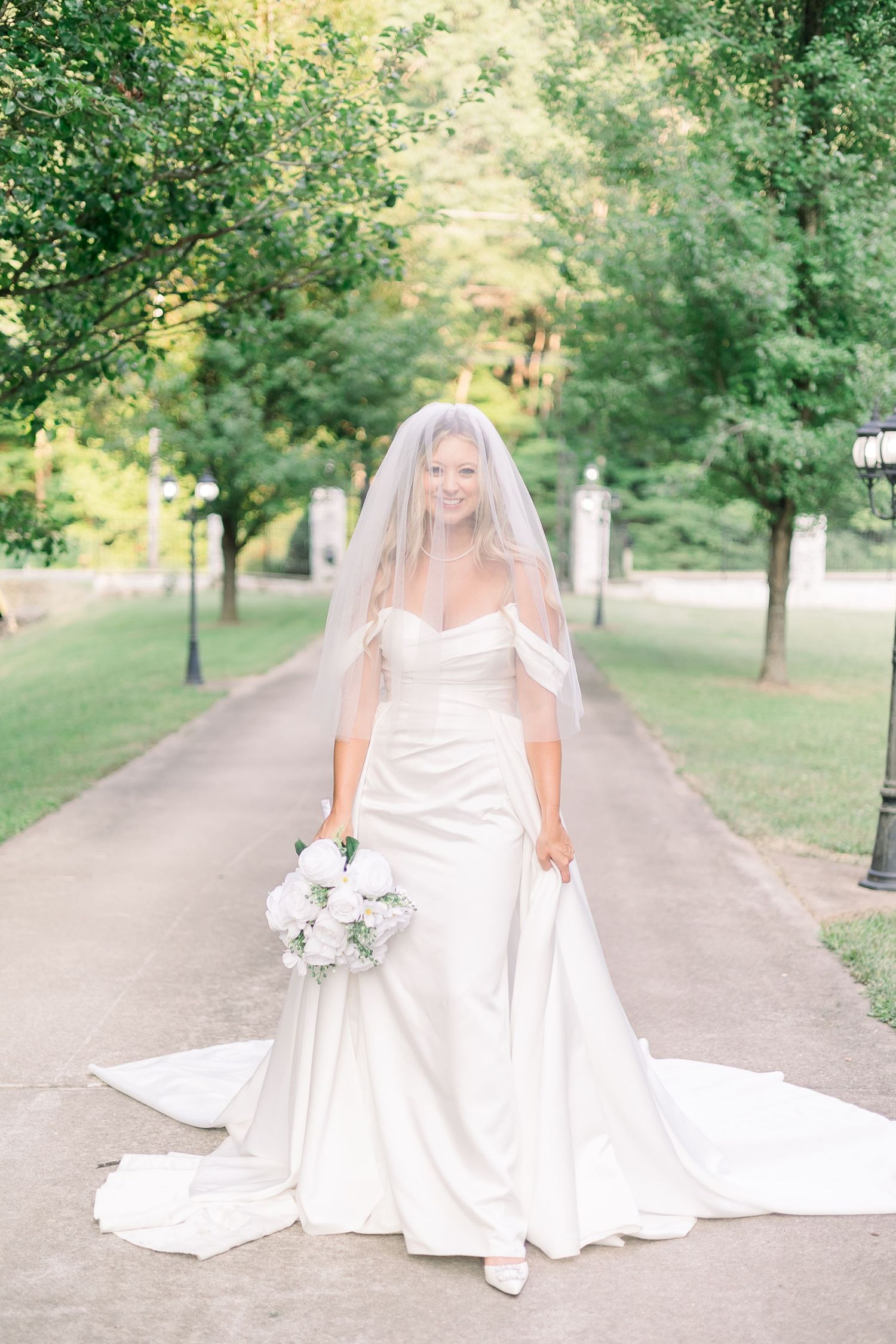 Bride in classic white gown poses gracefully on tree-lined garden path.