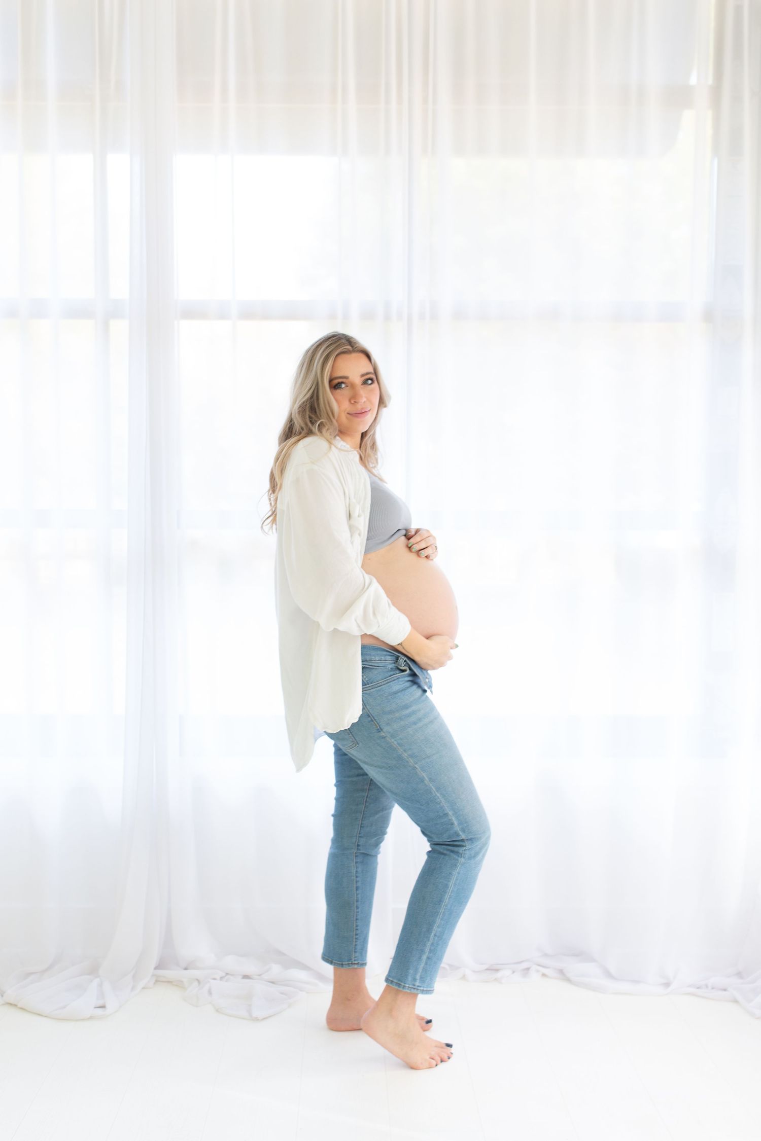 Expectant mother in casual attire poses sideways showing her baby bump against white curtained background.