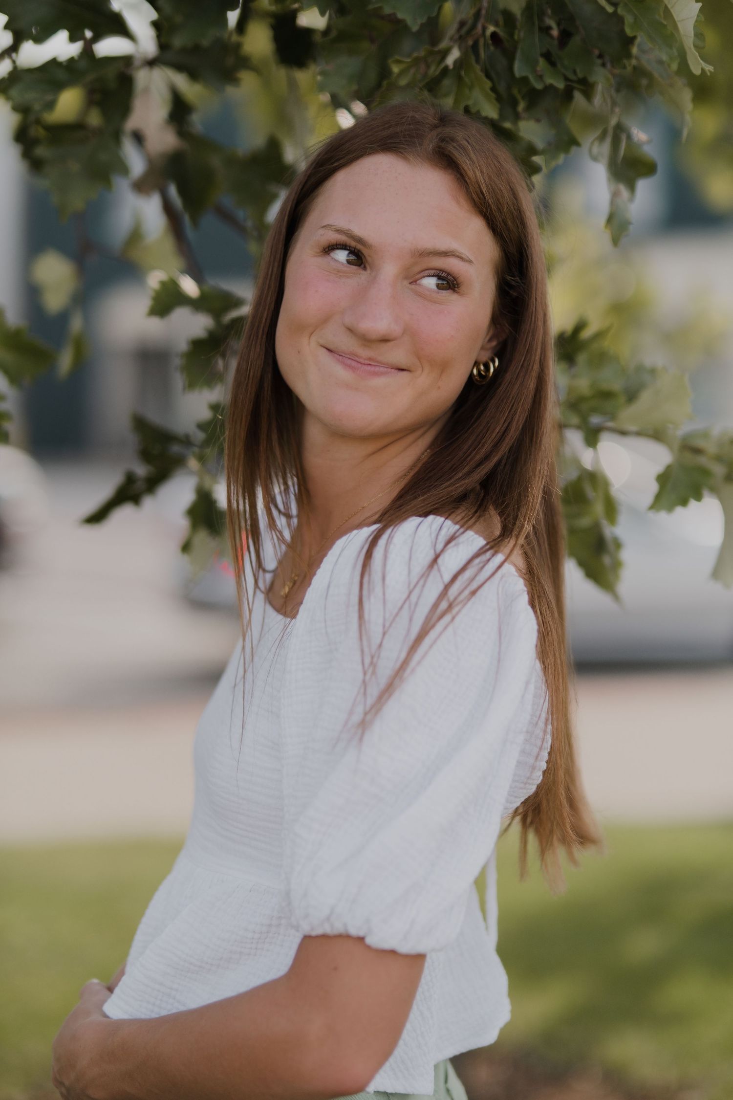 Someone in a white top poses playfully under green leaves in natural lighting.