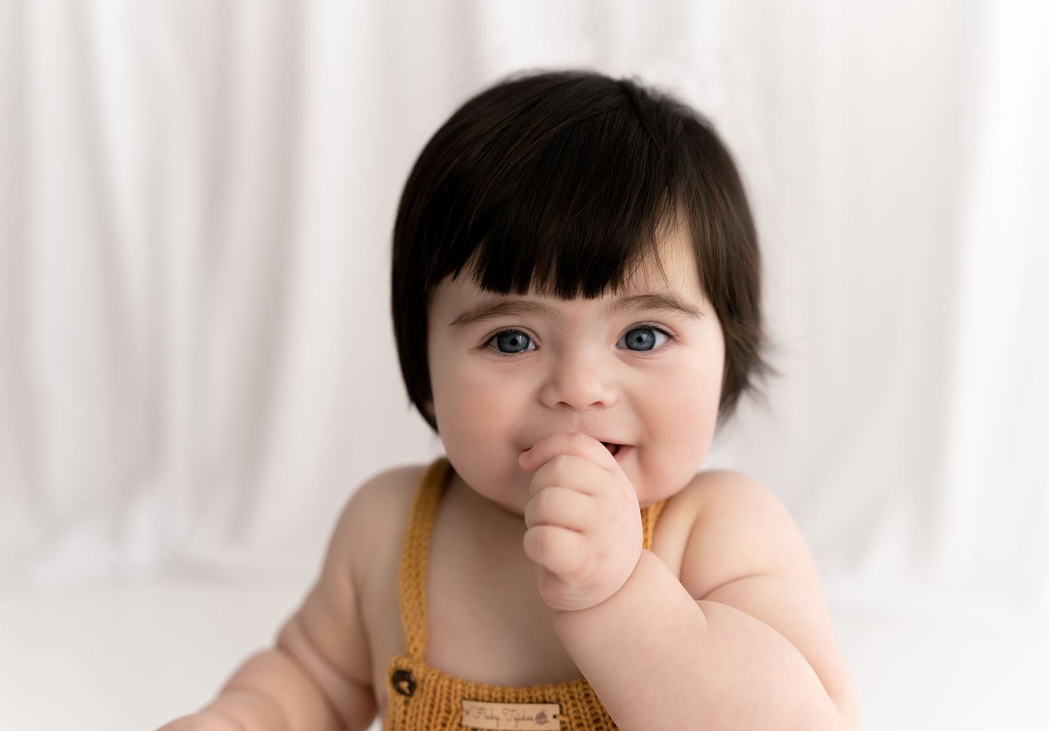 Baby in orange romper touches face while sitting against white backdrop.