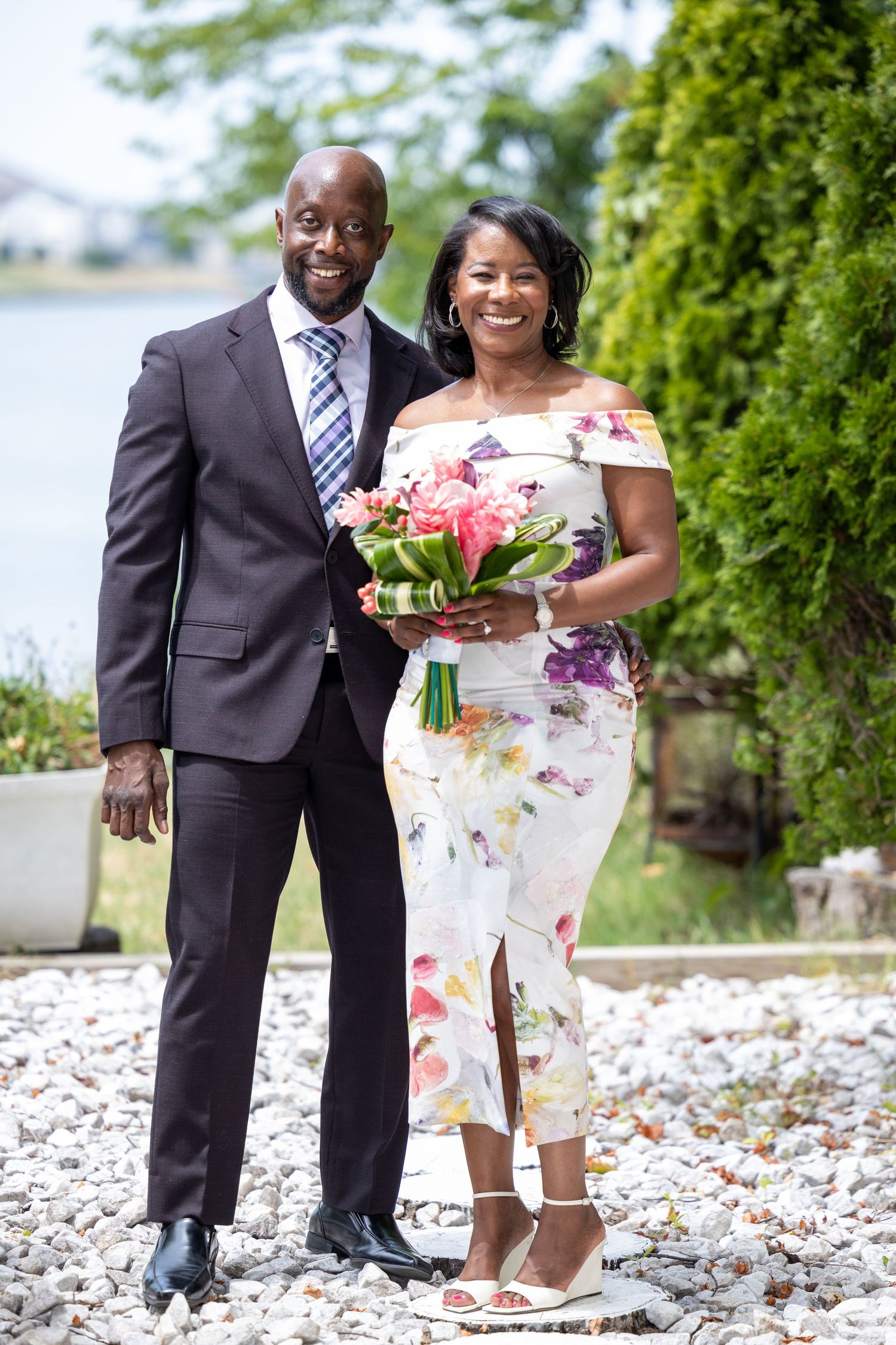 A couple poses outdoors in formal attire with a floral bouquet.