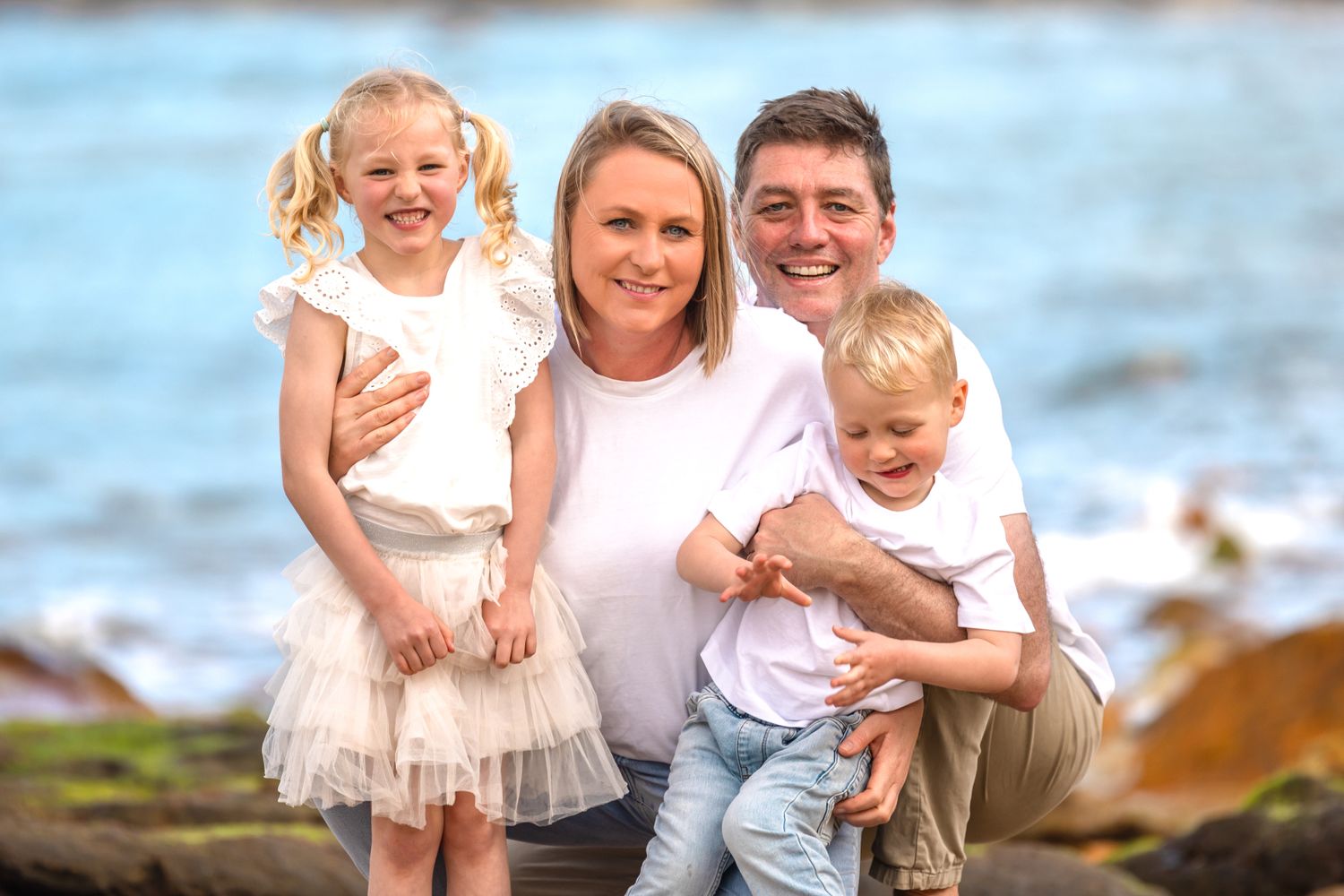 A smiling family of four, poses for a portrait outdoors with the ocean in the background.