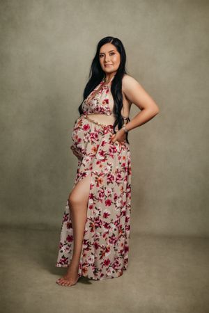 Pregnant woman in floral print dress poses against neutral backdrop for maternity photo.