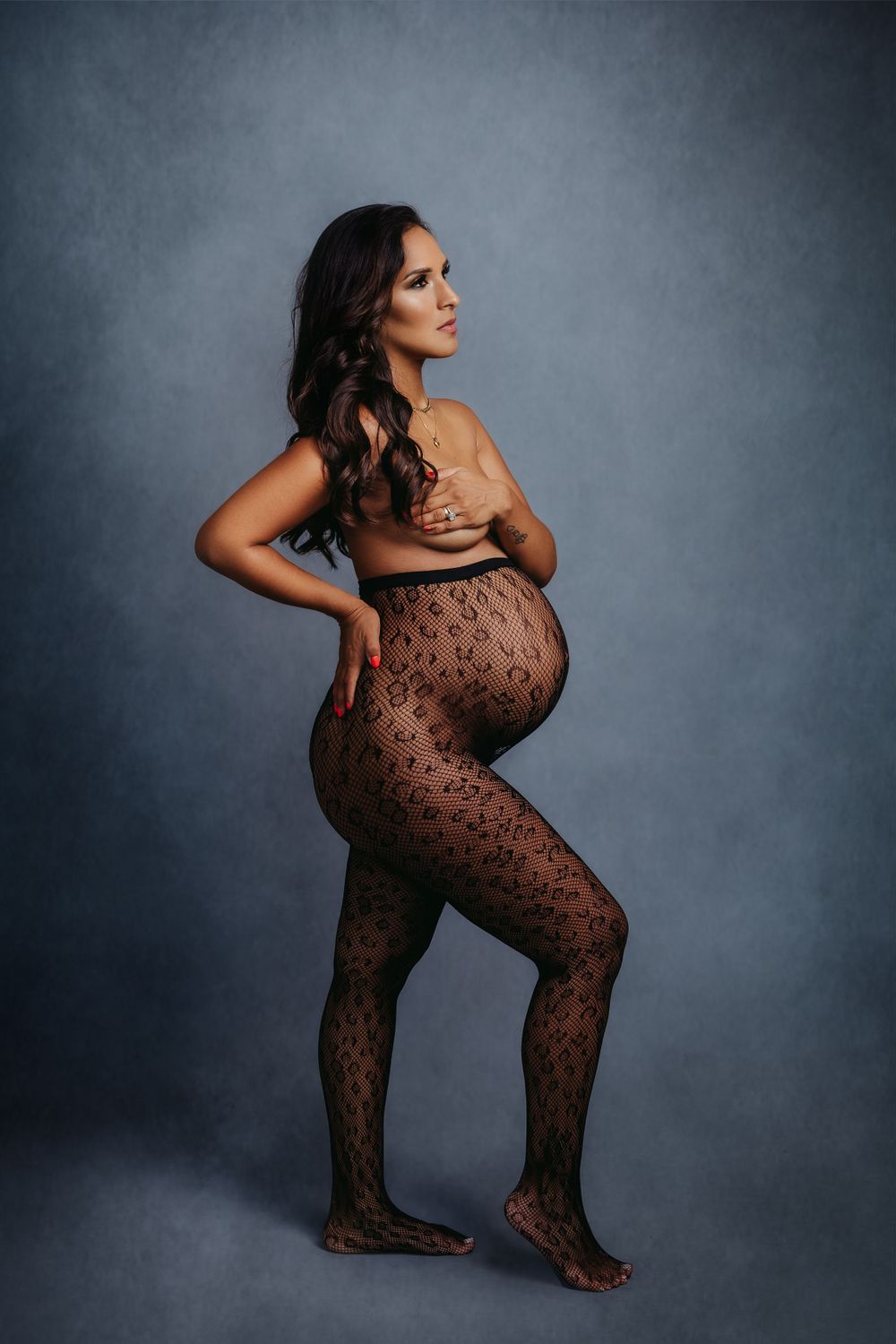 Side view of pregnant woman in lace bodysuit posing dramatically against gray backdrop.