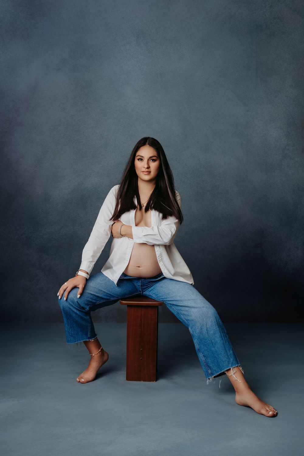 Woman in white shirt and jeans poses confidently on wooden stool during maternity session.