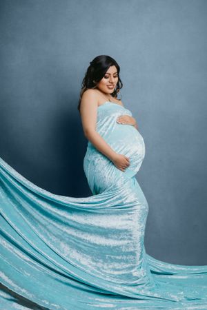 Maternity portrait featuring flowing aqua dress against blue studio background.
