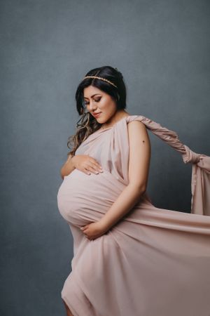 Elegant maternity portrait with flowing pink dress against gray backdrop.