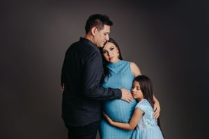 Family embraces pregnant mother in blue dress during intimate studio portrait session.