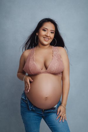 Pregnant woman in pink lace bralette and jeans poses against gray background for maternity photoshoot.