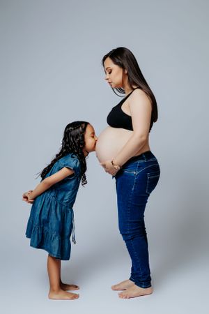 Young child interacts with pregnant mother's belly in studio portrait setting.