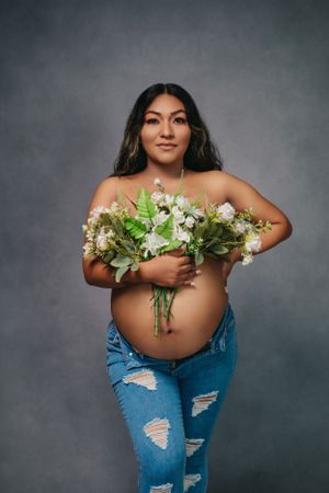 Pregnant woman in ripped jeans holds white floral bouquet against gray backdrop.