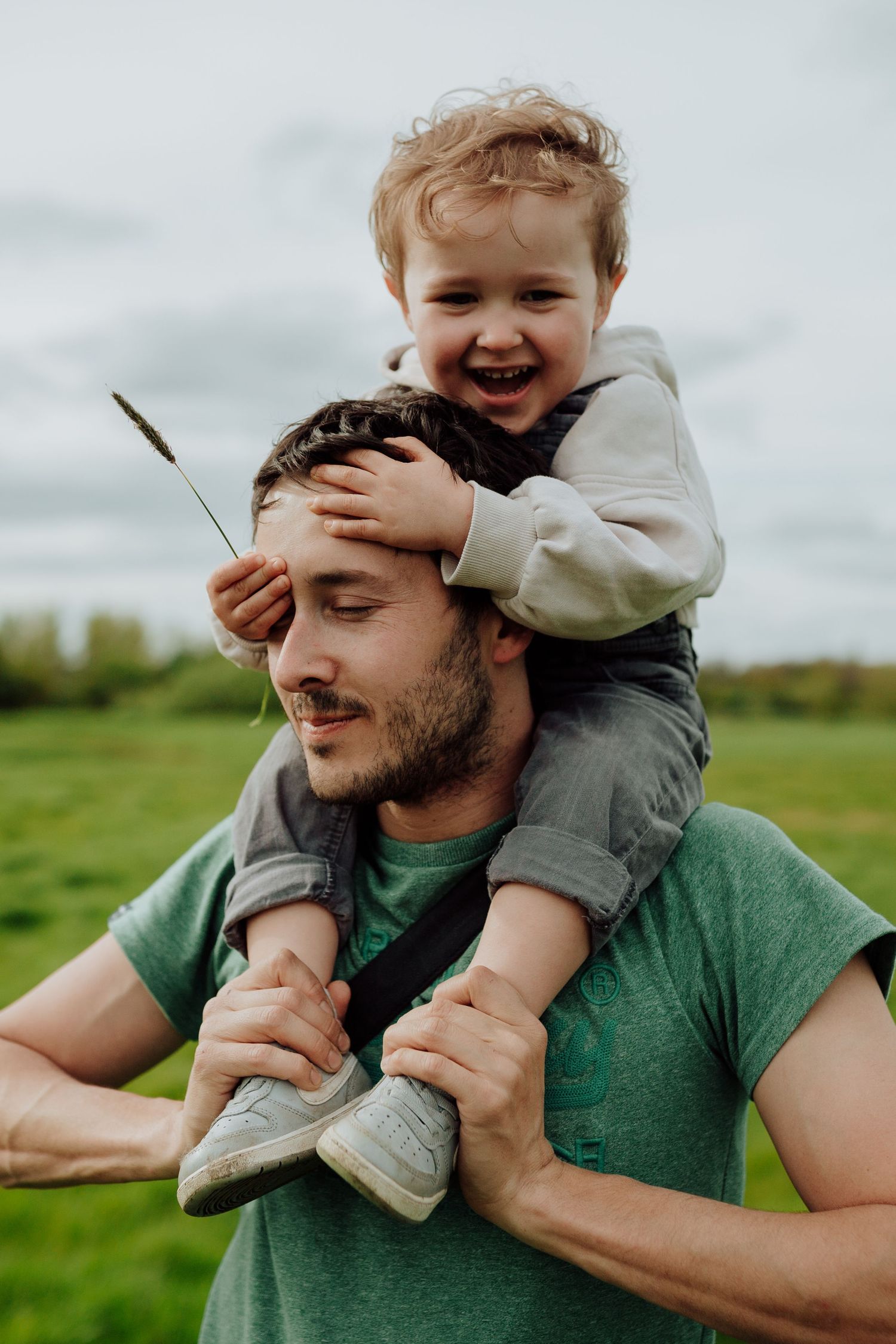 A touching moment of family fun as a laughing child rides on shoulders in a green field on a cloudy day.