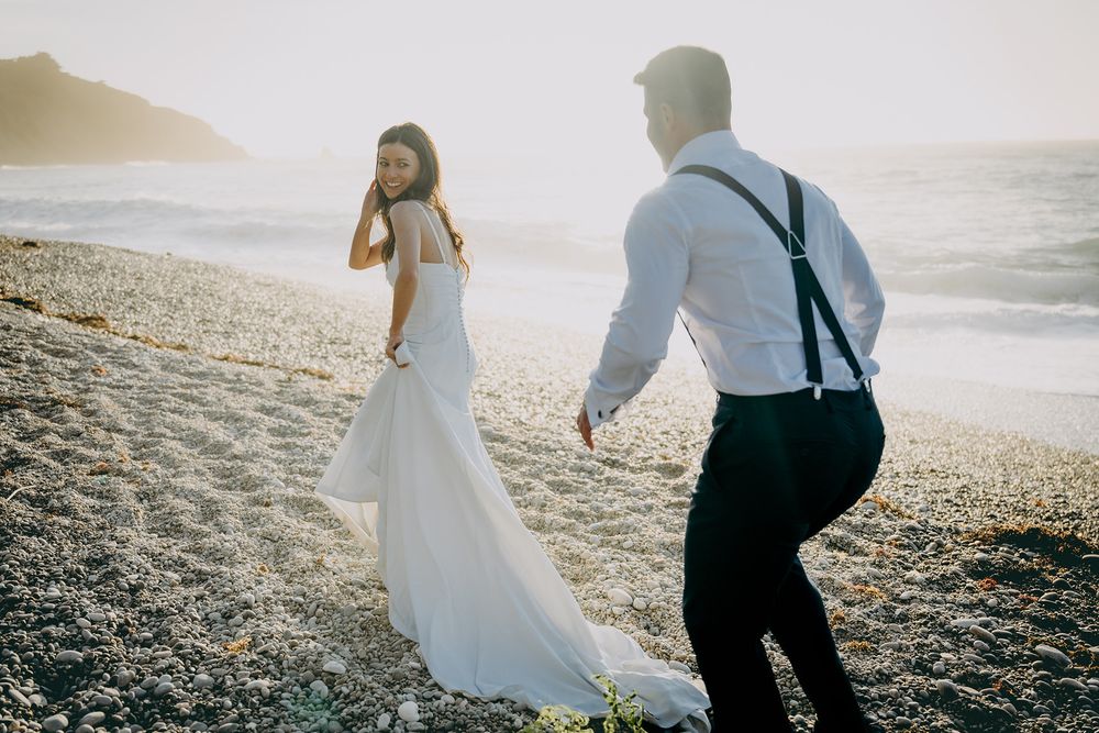 Pareja de recién casados caminando por una playa de Asturias al atardecer durante una sesión de postboda fotografiada por Juan Llavio