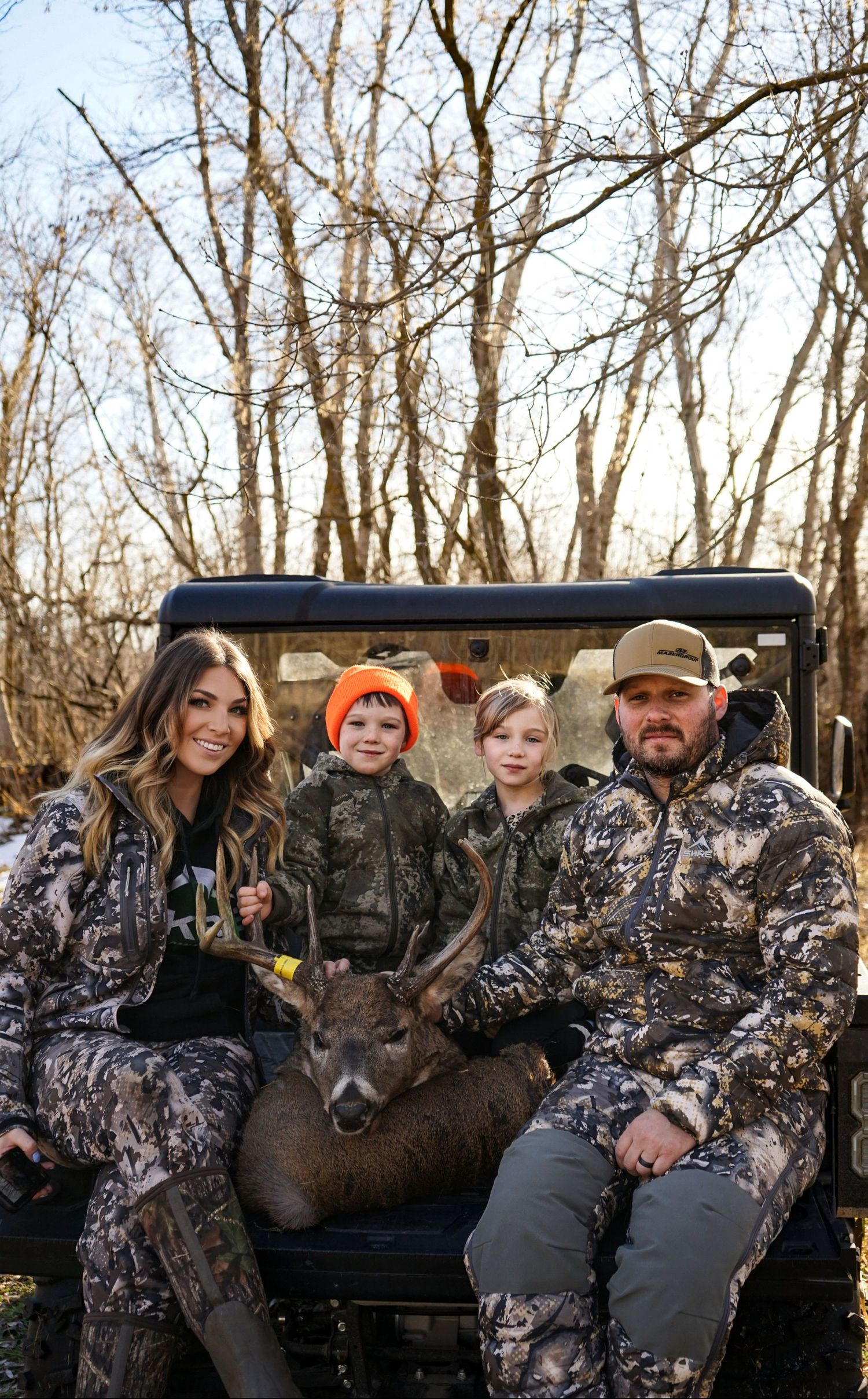 A family in camouflage hunting gear poses with their harvested deer in a utility vehicle.