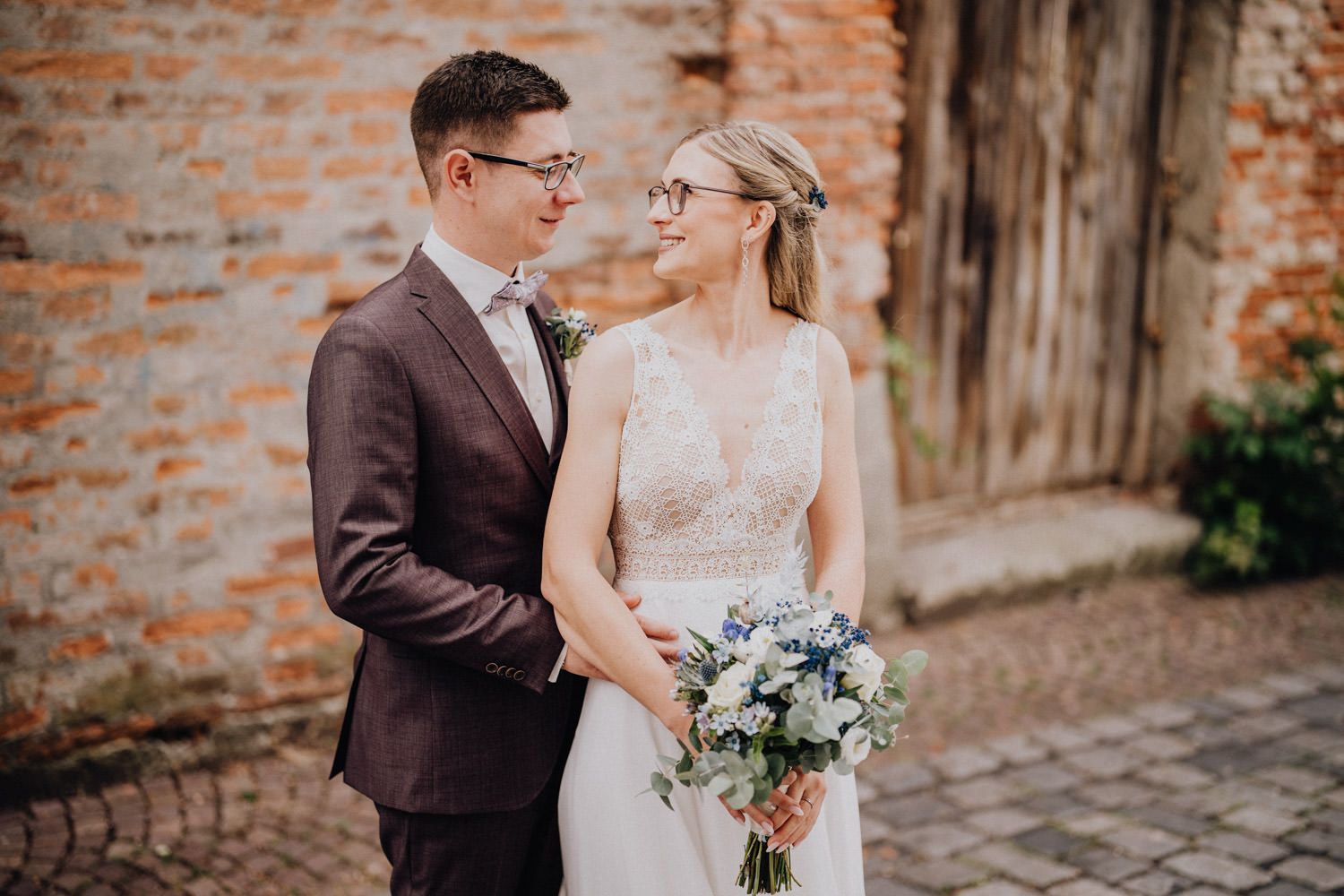 Elegantly dressed couple sharing an intimate moment against a rustic brick wall with blue floral bouquet.