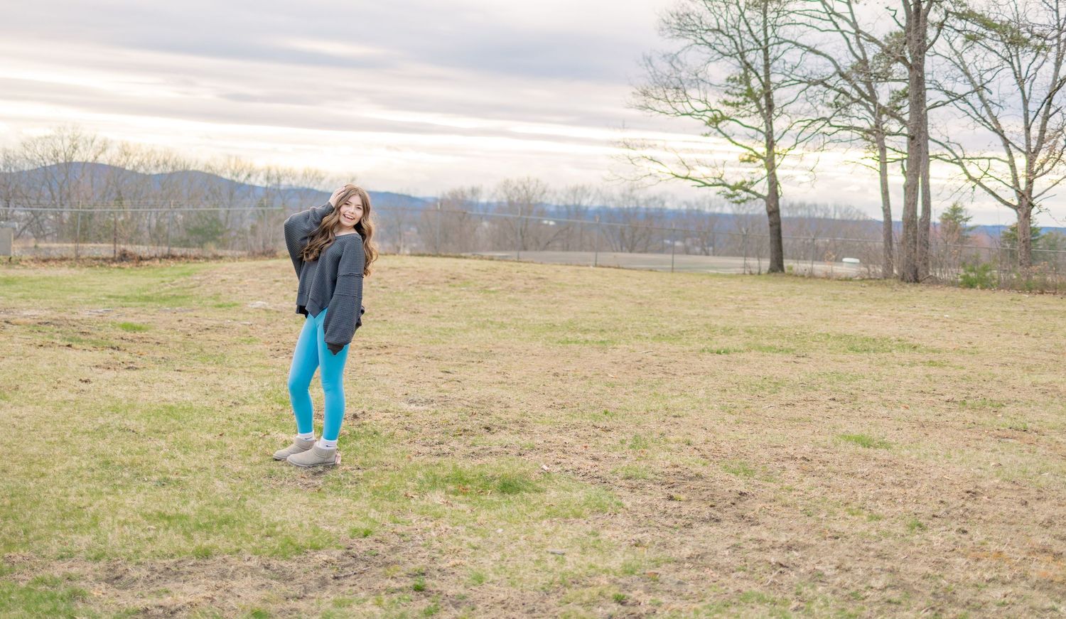 Figure in navy sweater and blue leggings stands in a scenic field with trees in background.