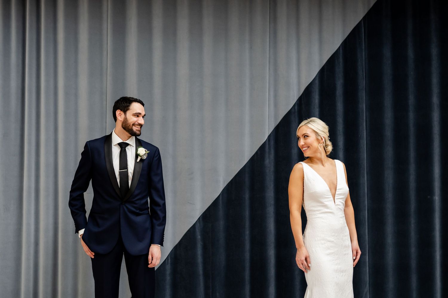 Bride in a white gown and groom in a navy suit with a boutonniere smile at each other against a split gray and dark curtain backdrop.