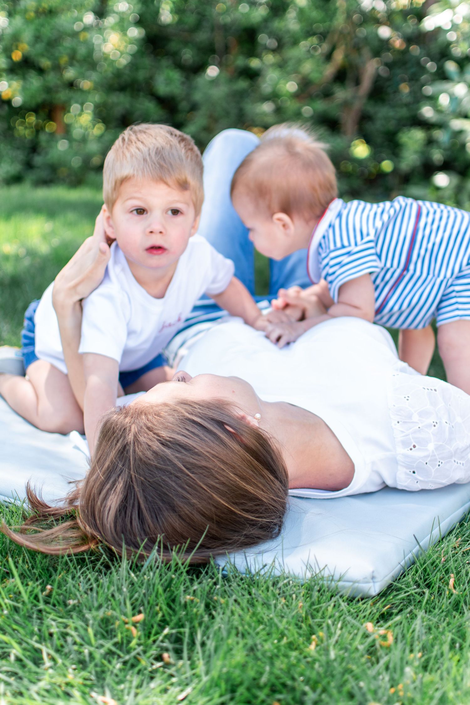 Two boys climb on top of their mom as they relax on a blanket in the back yard.