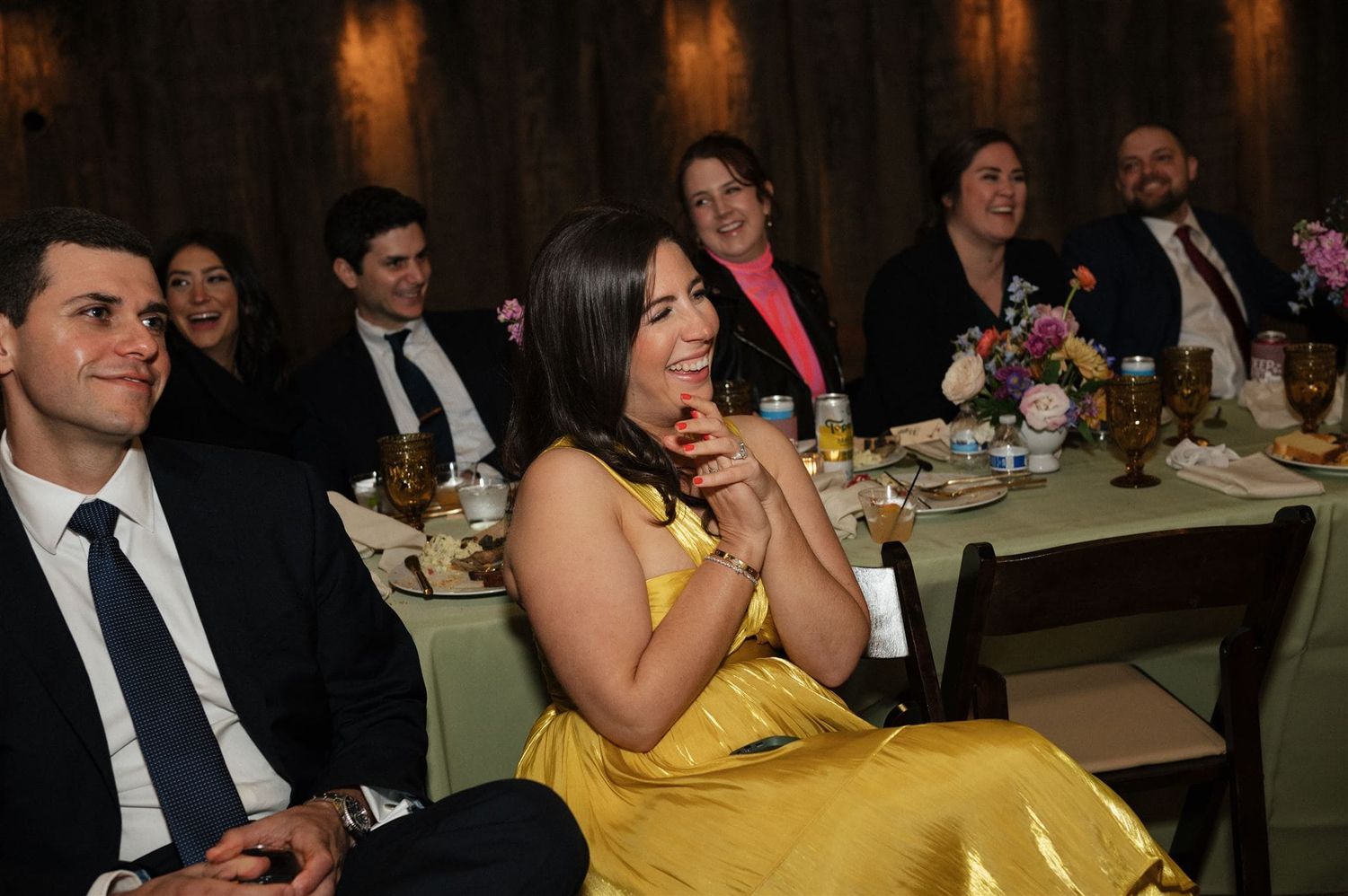 Wedding guests seated at dinner tables during an evening reception share laughter and smiles.