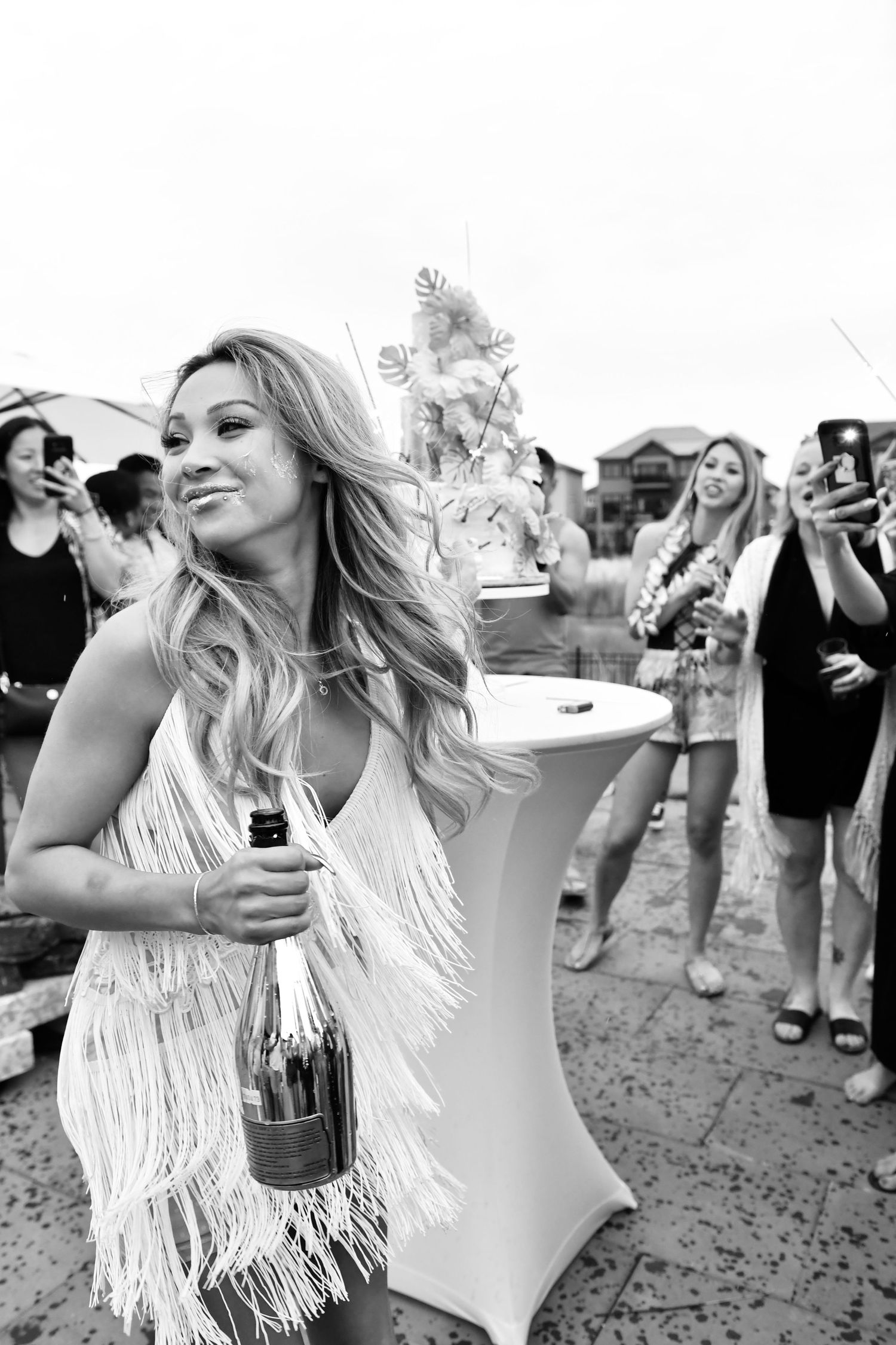 A woman in a white fringed dress dances at an outdoor festival while holding a bottle of champagne.