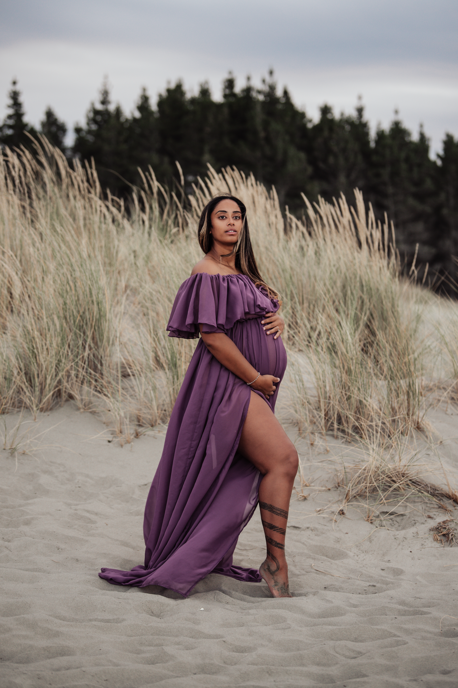 A purple off-shoulder maternity gown flows gracefully against beach dunes and evergreen trees.