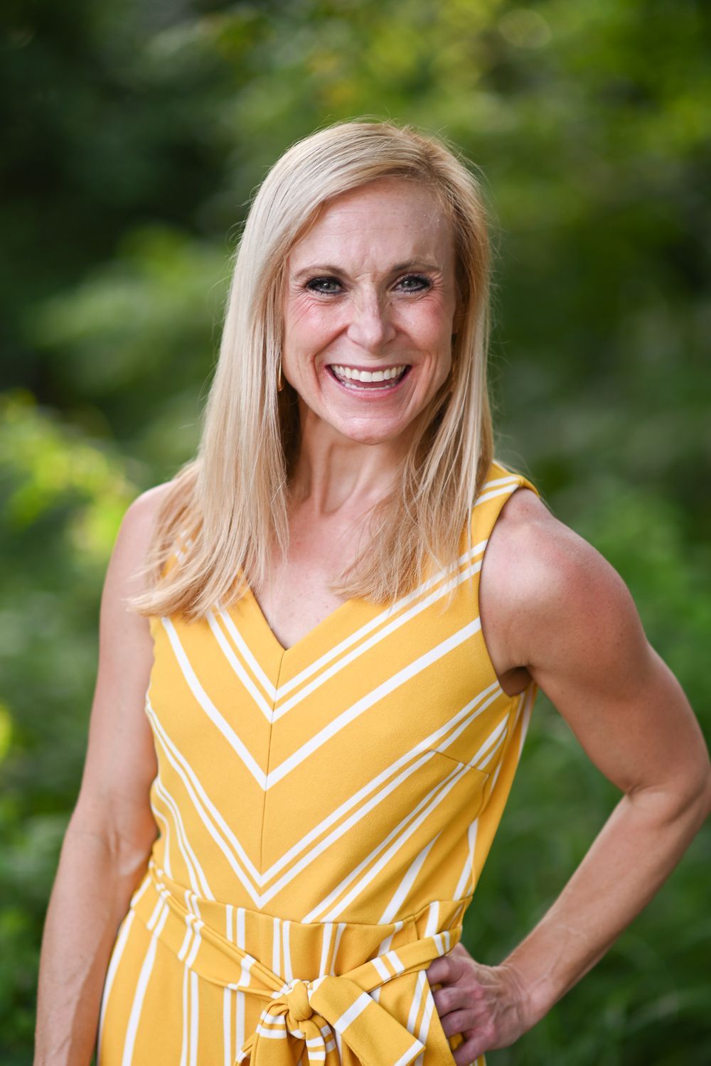 Smiling blonde woman in yellow striped sleeveless dress posing outdoors with green foliage background.