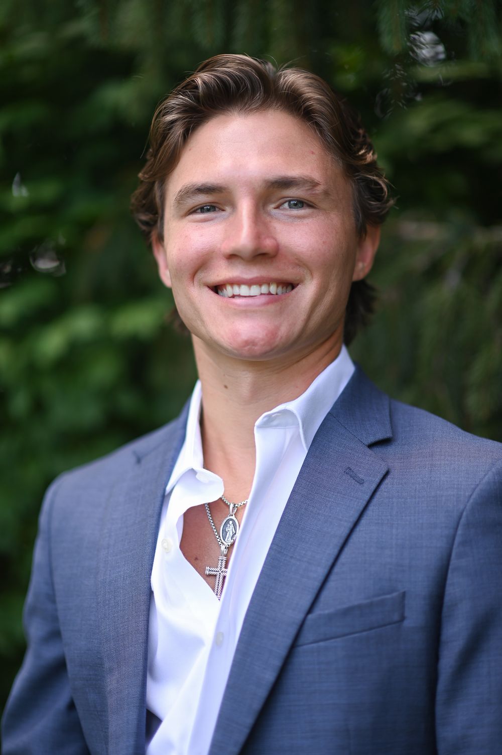 Smiling young man in gray suit and white shirt wearing a cross necklace, posing outdoors with green trees behind.