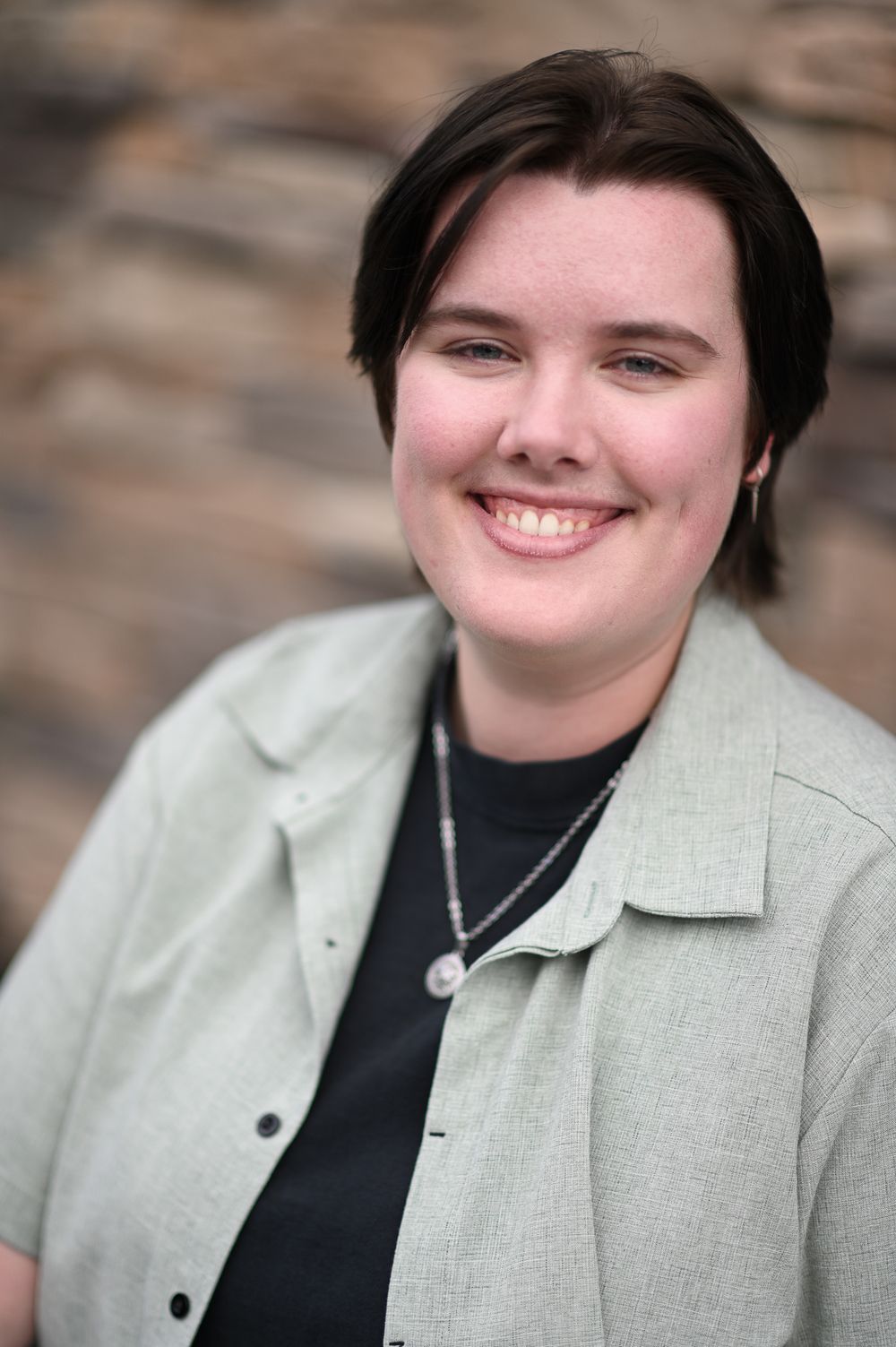 Smiling young woman with short dark hair wearing a light gray blazer and black shirt outdoors.