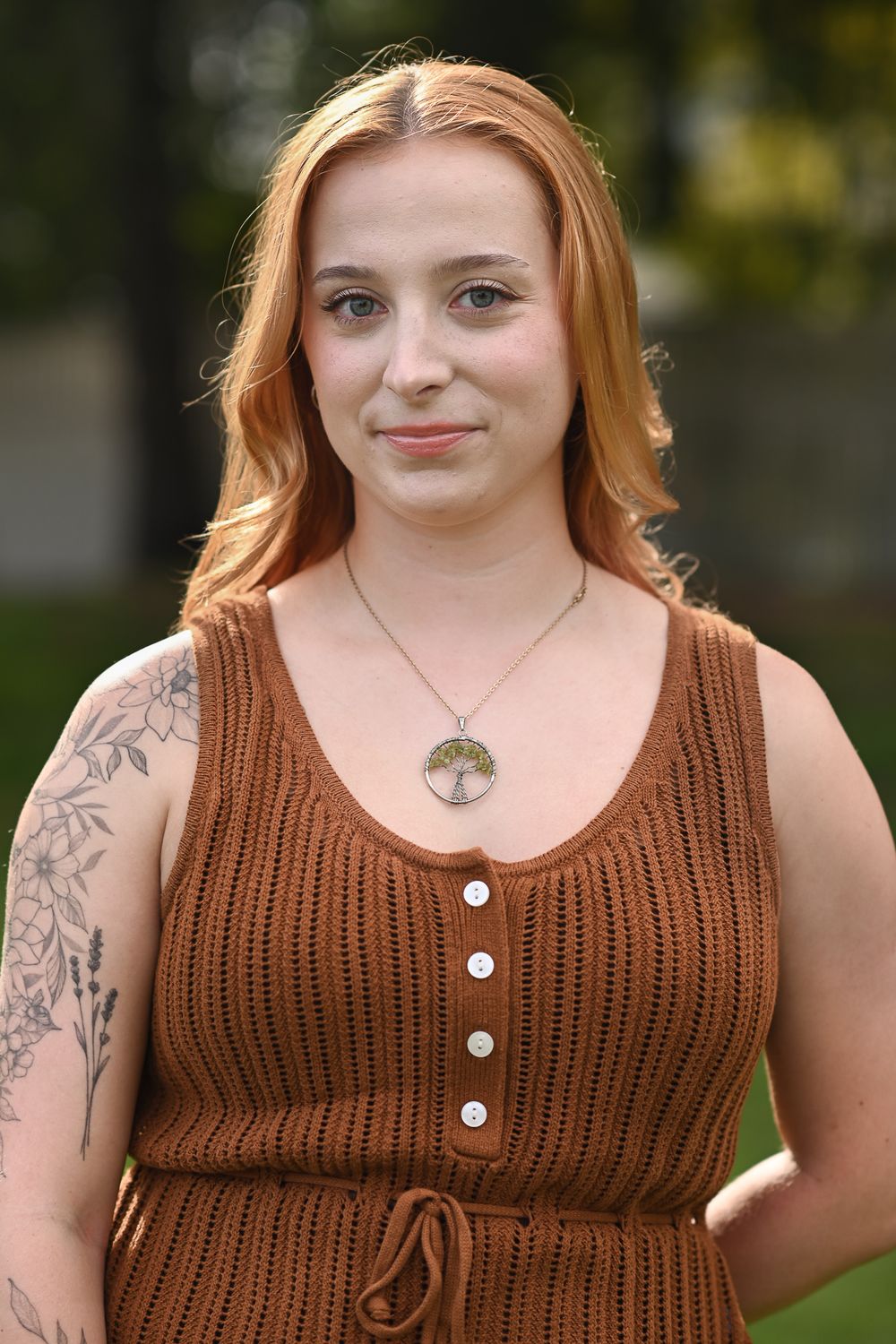 Young woman with red hair wearing a brown knit dress and tree of life necklace, with floral arm tattoo, outdoors.