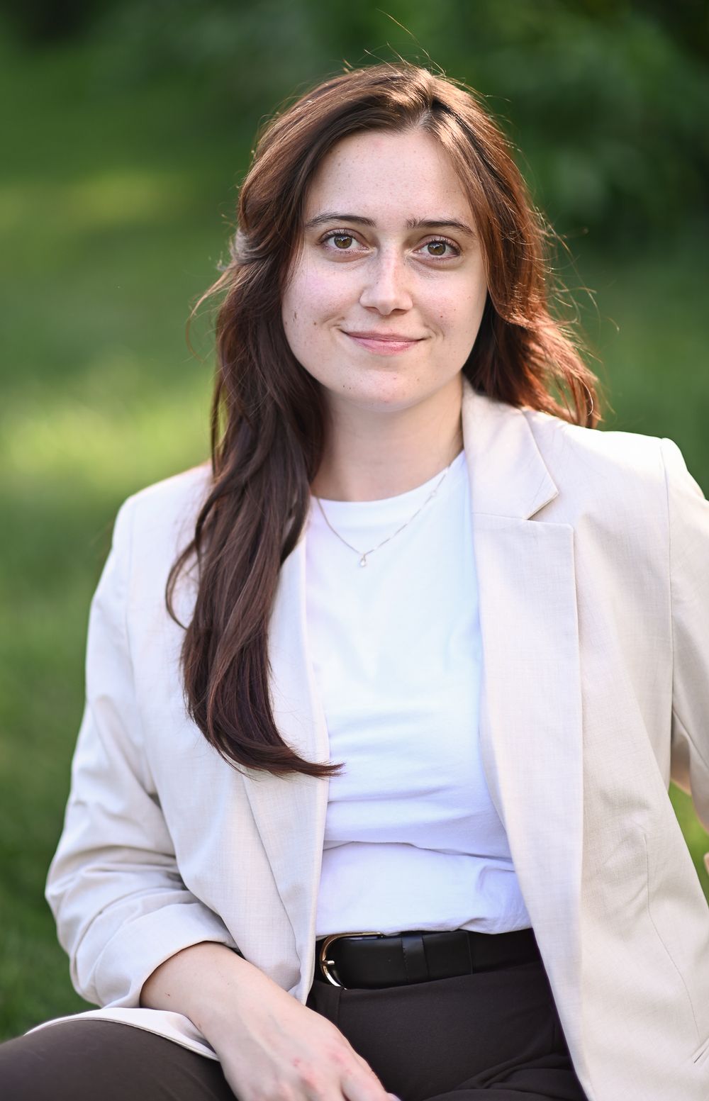 Young woman with long brown hair in white blazer smiling outdoors in a green park setting.