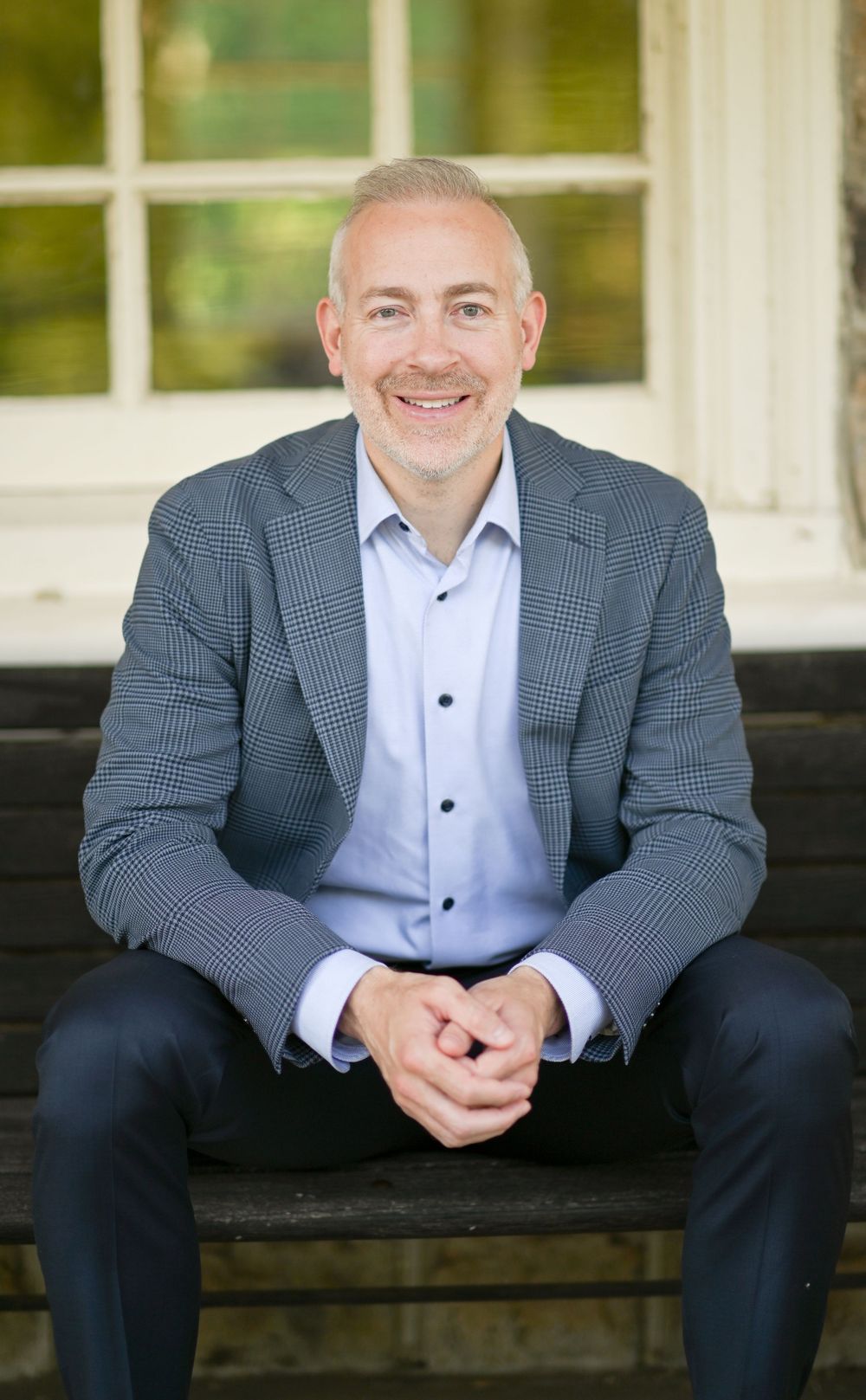 Smiling man in gray blazer and light blue shirt sits on bench outdoors, hands clasped, in a professional headshot.