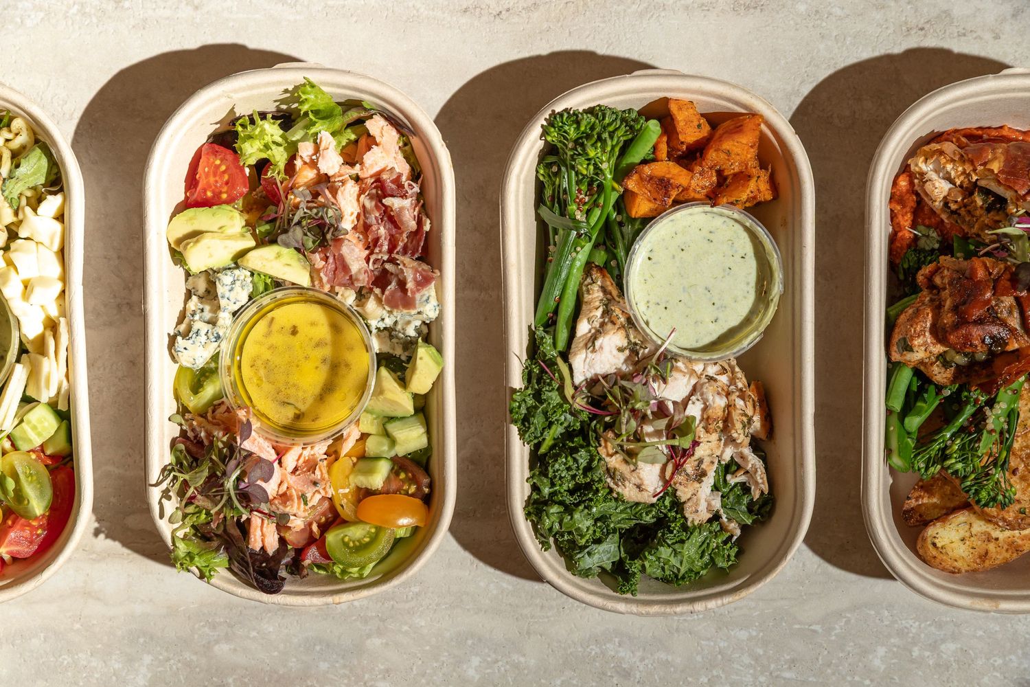 Four oval meal prep containers with fresh salads, roasted vegetables, grilled chicken, and dressings on a light surface.