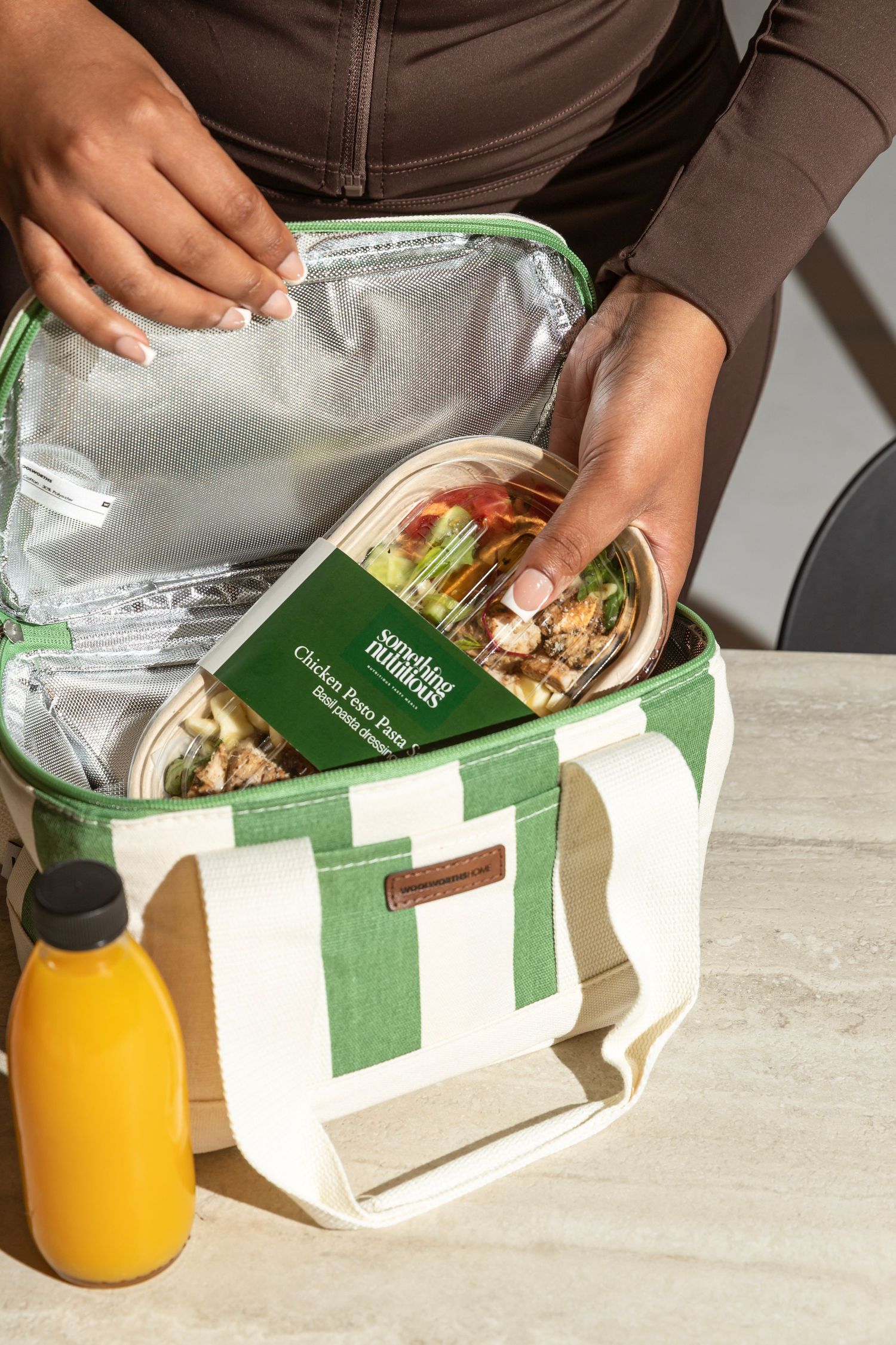 Woman placing a Cooking Matters meal kit into a insulated green striped lunch bag beside an orange juice bottle.