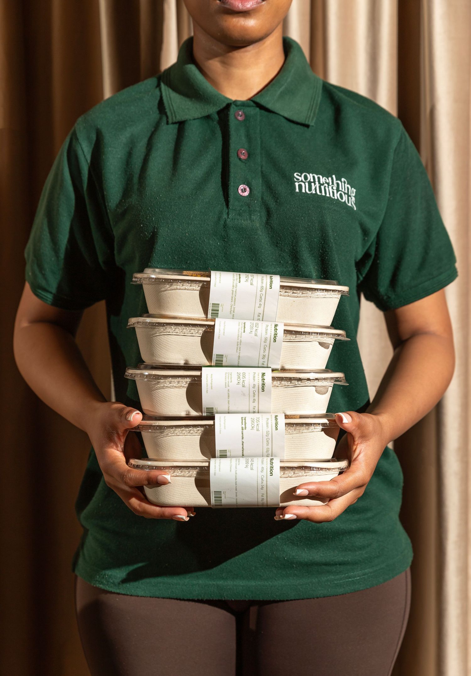 Person in green 'Something Nutritious' polo shirt holding stacked food containers with labels.