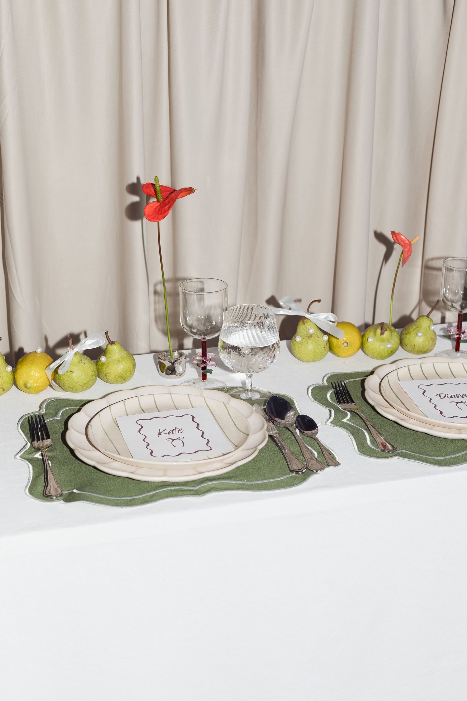 Elegant table setting with green placemats, white plates, fresh apples and red flower accents against a light curtain backdrop.