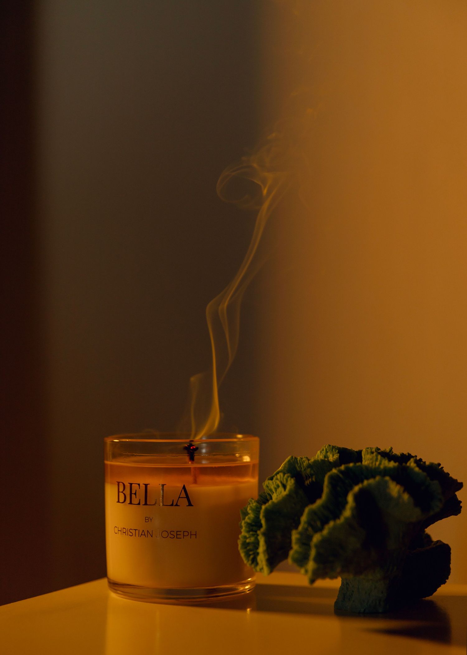 Candle with 'BELLA' text label sits beside fresh green kale leaves against a dark background in warm lighting.