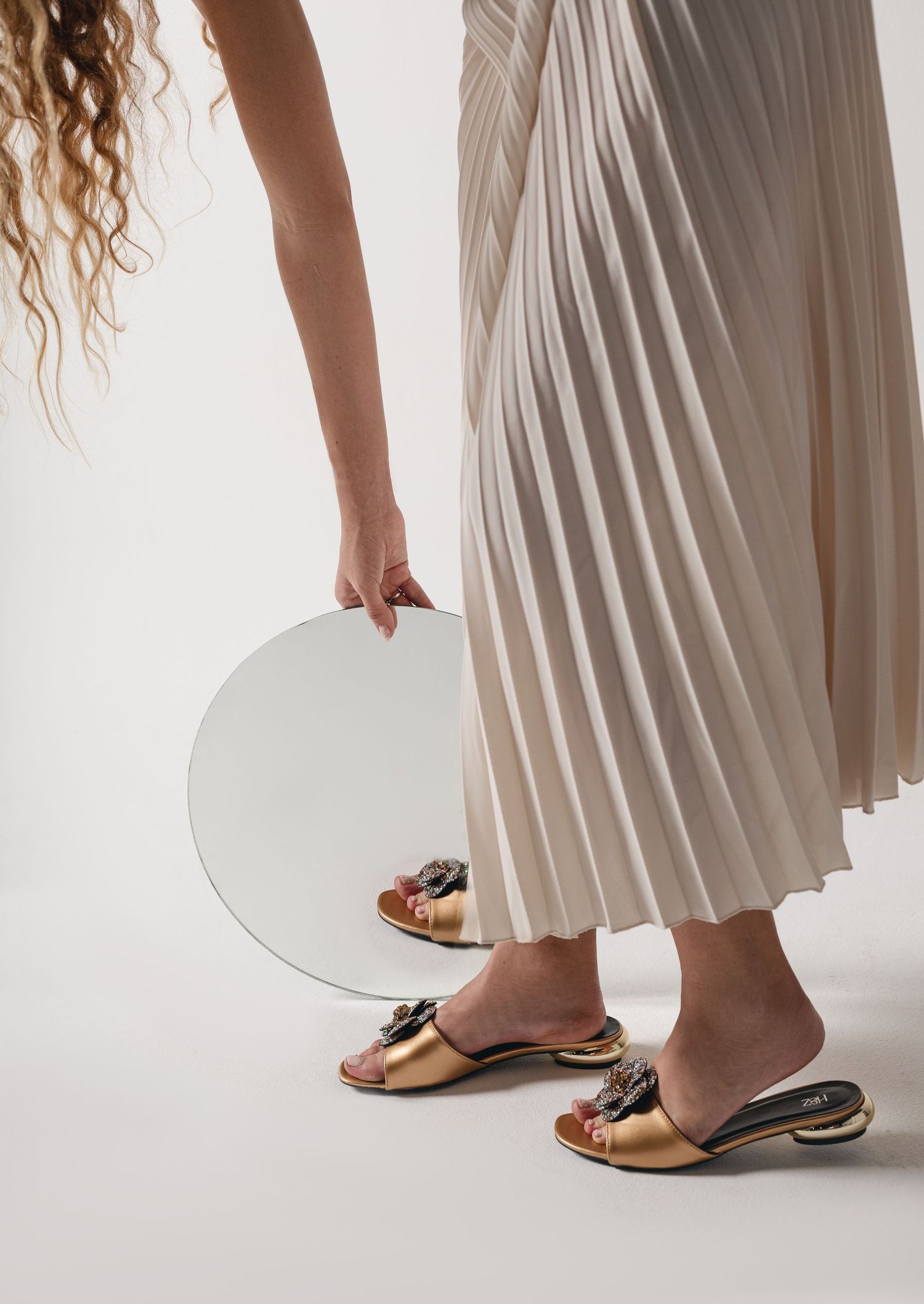 Elegant gold sandals and a flowing pleated skirt shown with a circular mirror in a minimalist fashion setting.