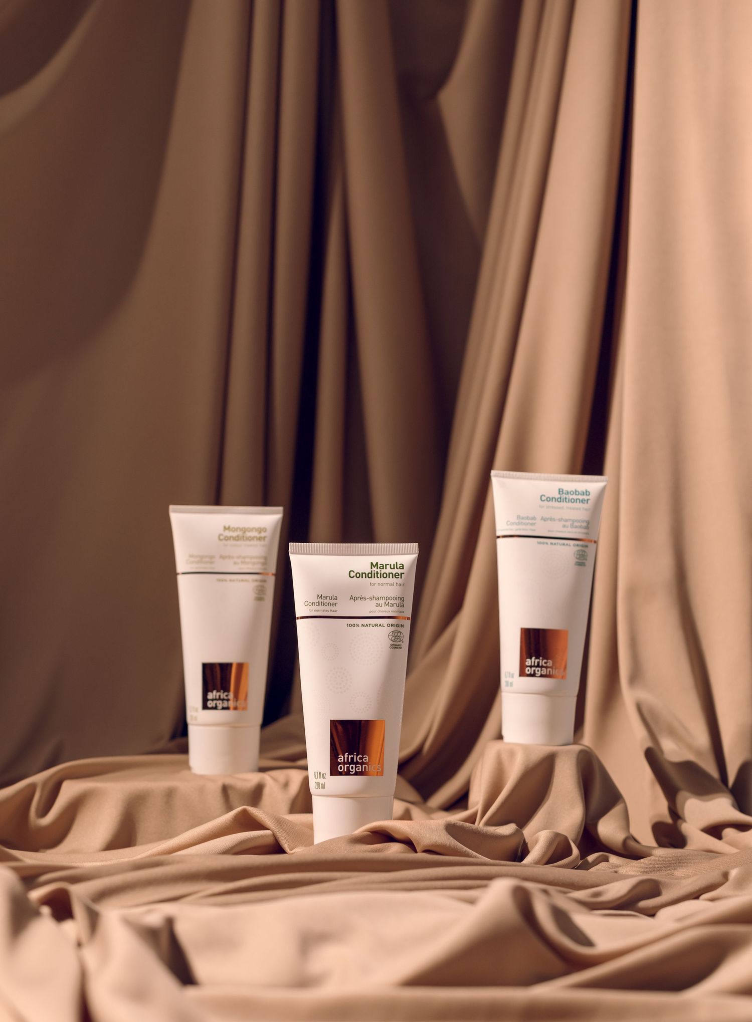 Three skincare product tubes arranged on flowing beige satin fabric against a neutral brown background.