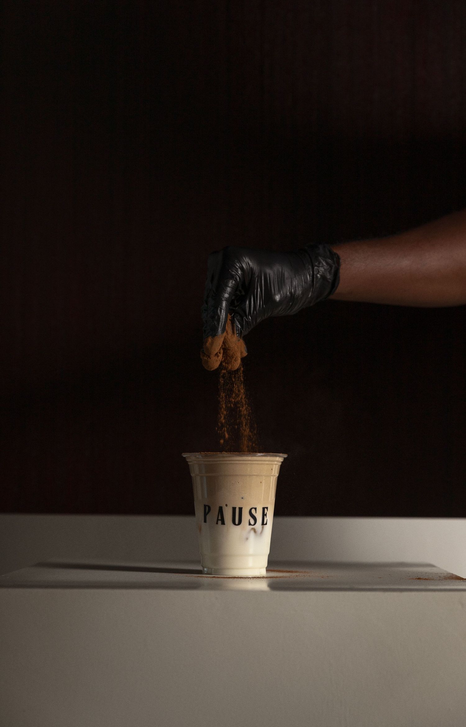 A gloved hand crumbles ingredients into a Pause branded cup on a white surface against a dark background.