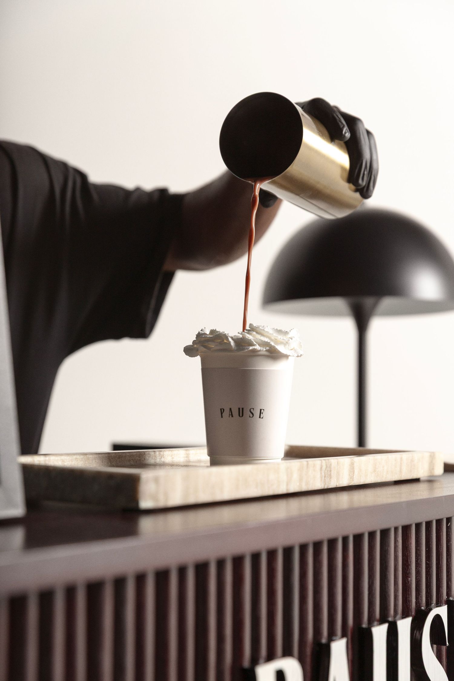 Barista pouring chocolate sauce into a whipped cream topped cup labeled PAUSE on a wooden tray beside a black lamp.