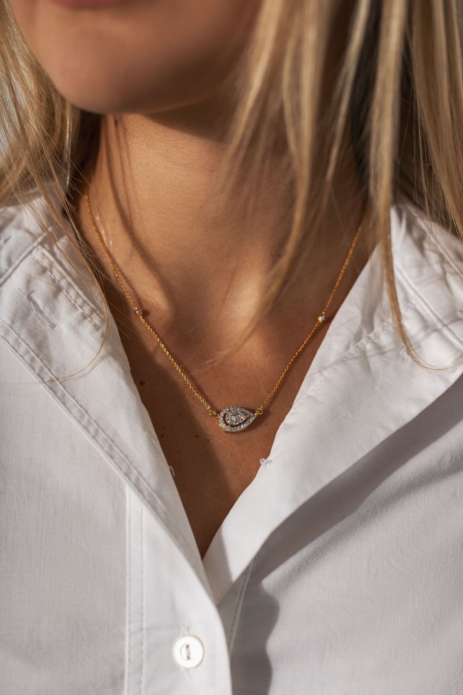 Delicate gold necklace with sparkly eye-shaped pendant worn with crisp white button-down shirt in sunlight.
