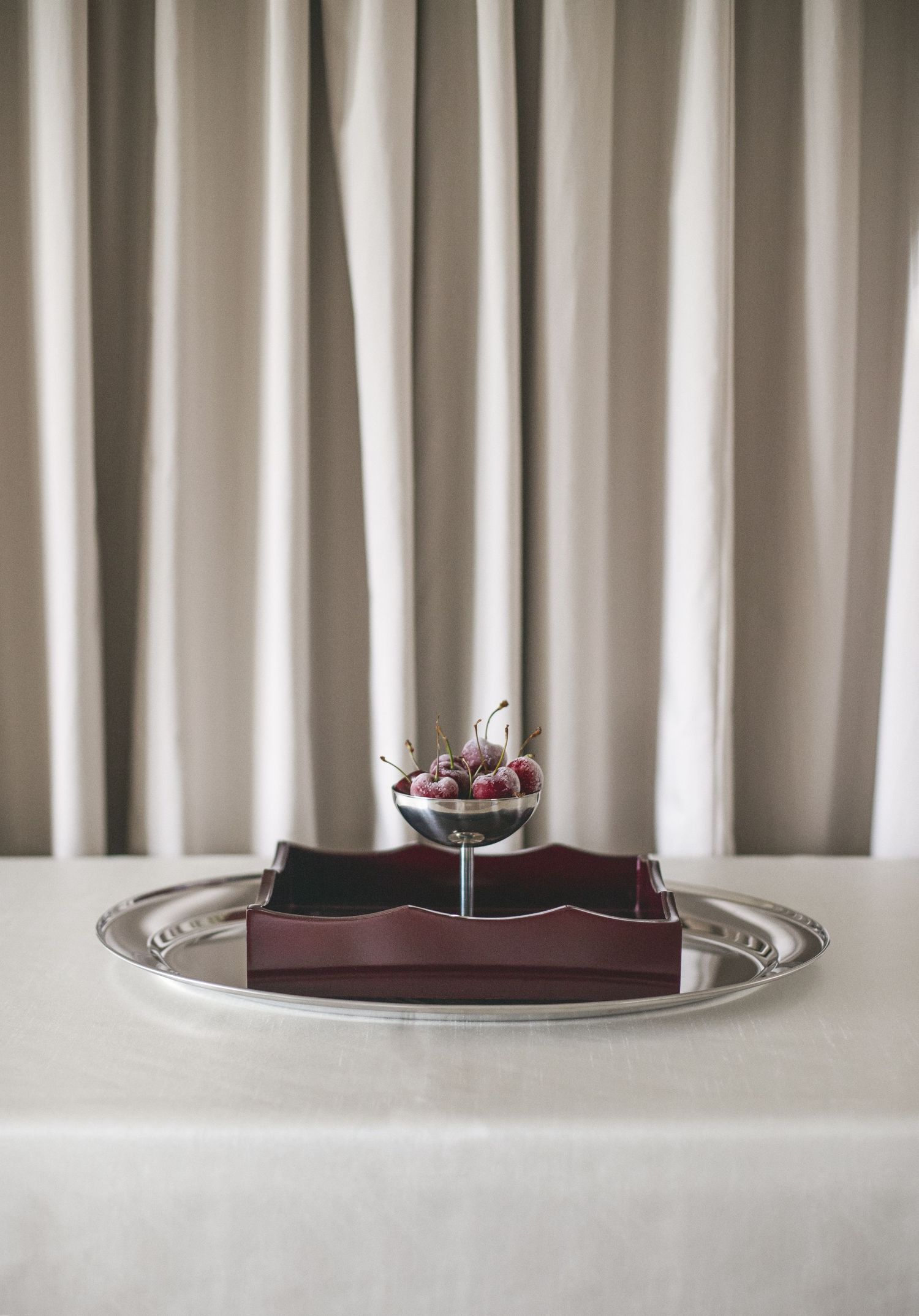 A silver tray holds a leather portfolio and small glass bowl with berries on a white tablecloth against draped curtains.