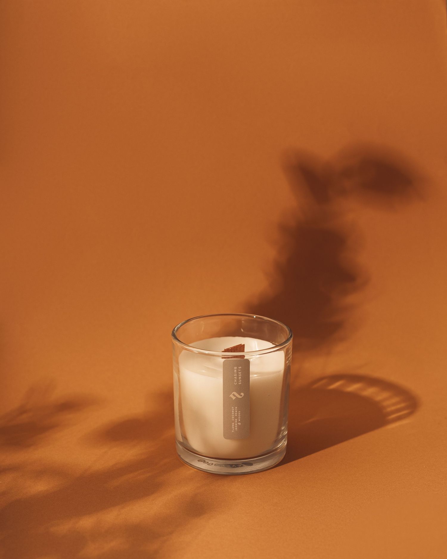 A glowing white candle in a clear glass vessel casts dramatic shadows against a warm terracotta-colored background.