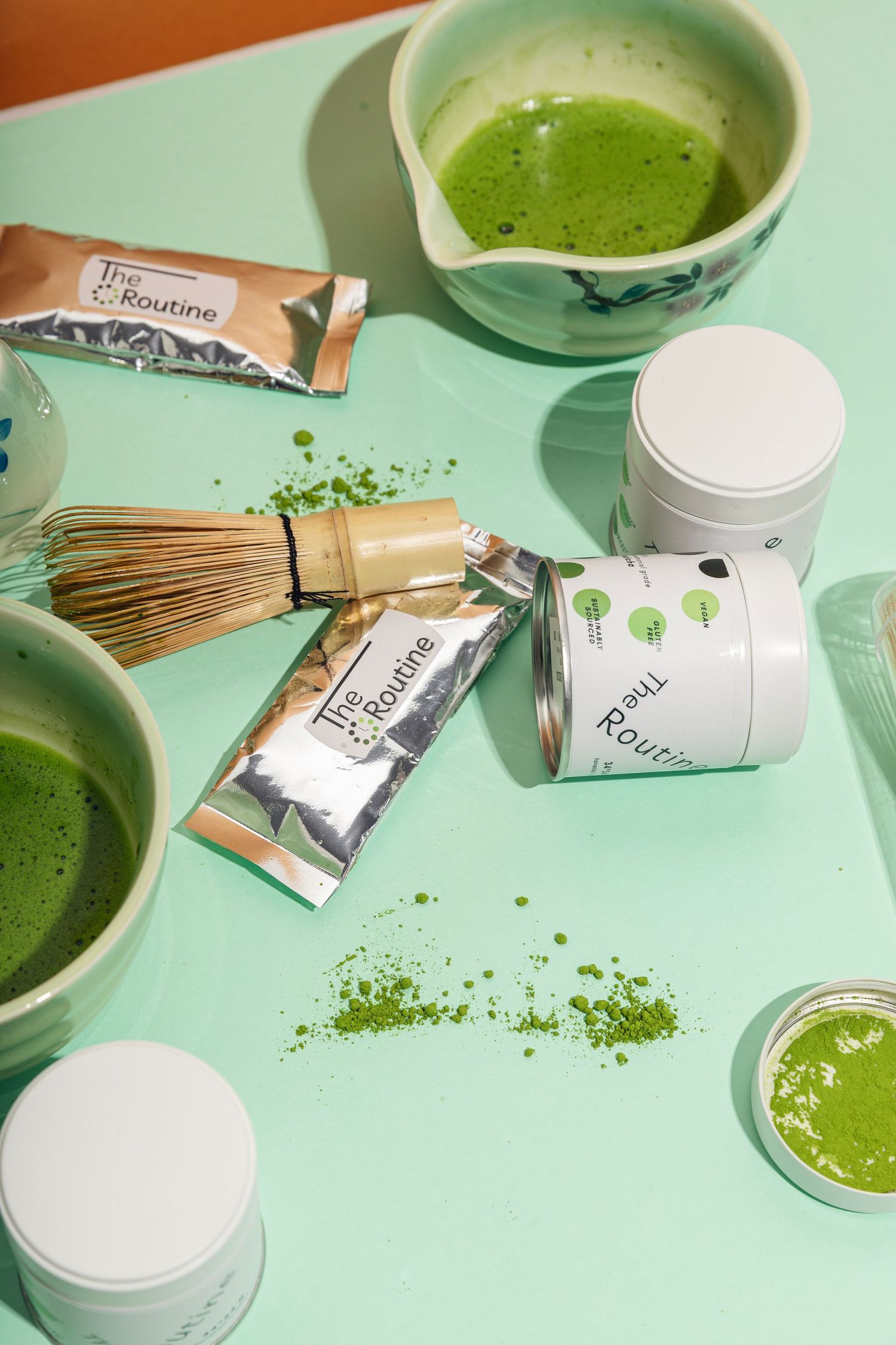 Green matcha tea ceremony setup with bamboo whisk, traditional bowls and modern powder containers on mint green surface.
