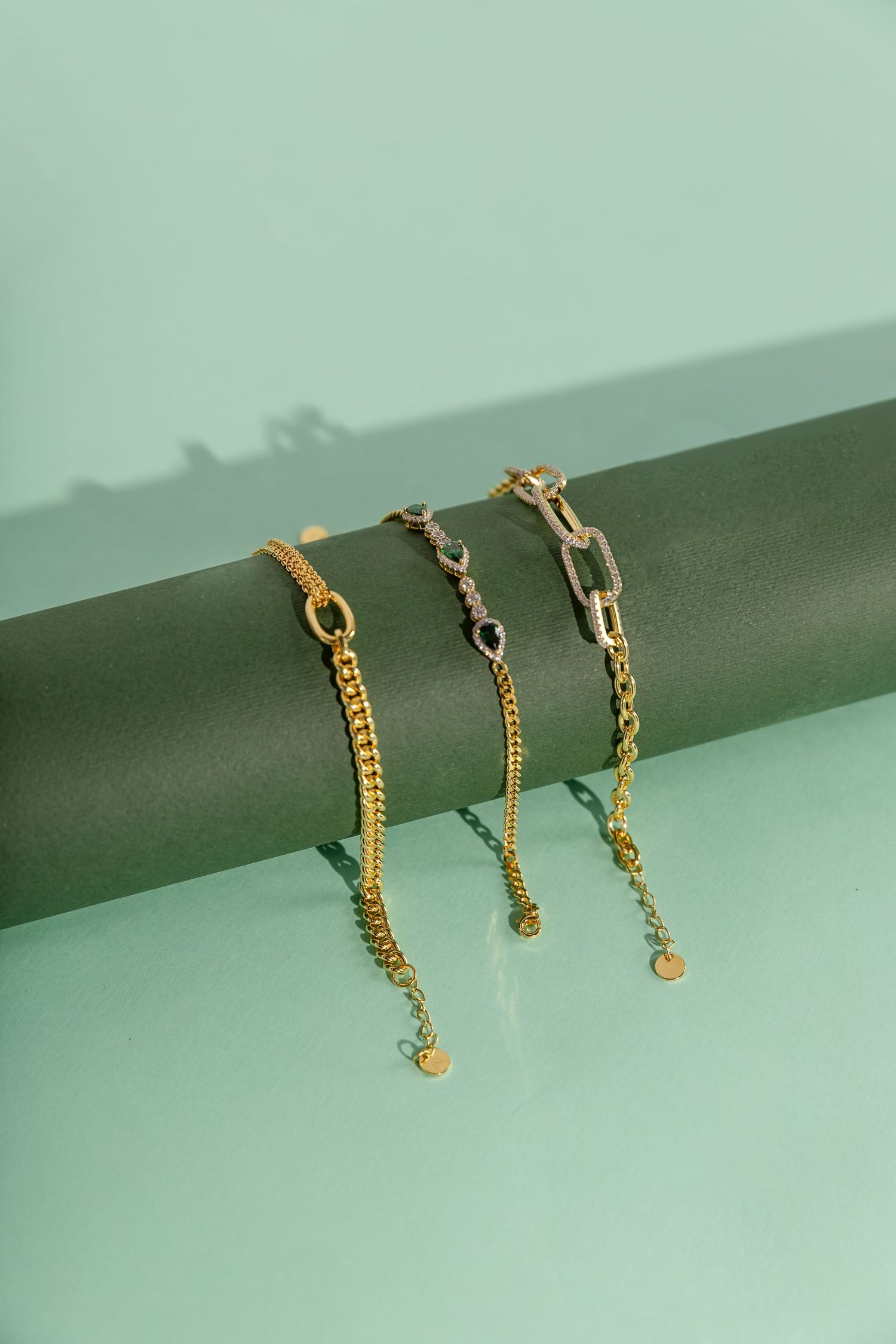 Three delicate gold chain bracelets with various link designs displayed on a grey cylinder against mint green background.