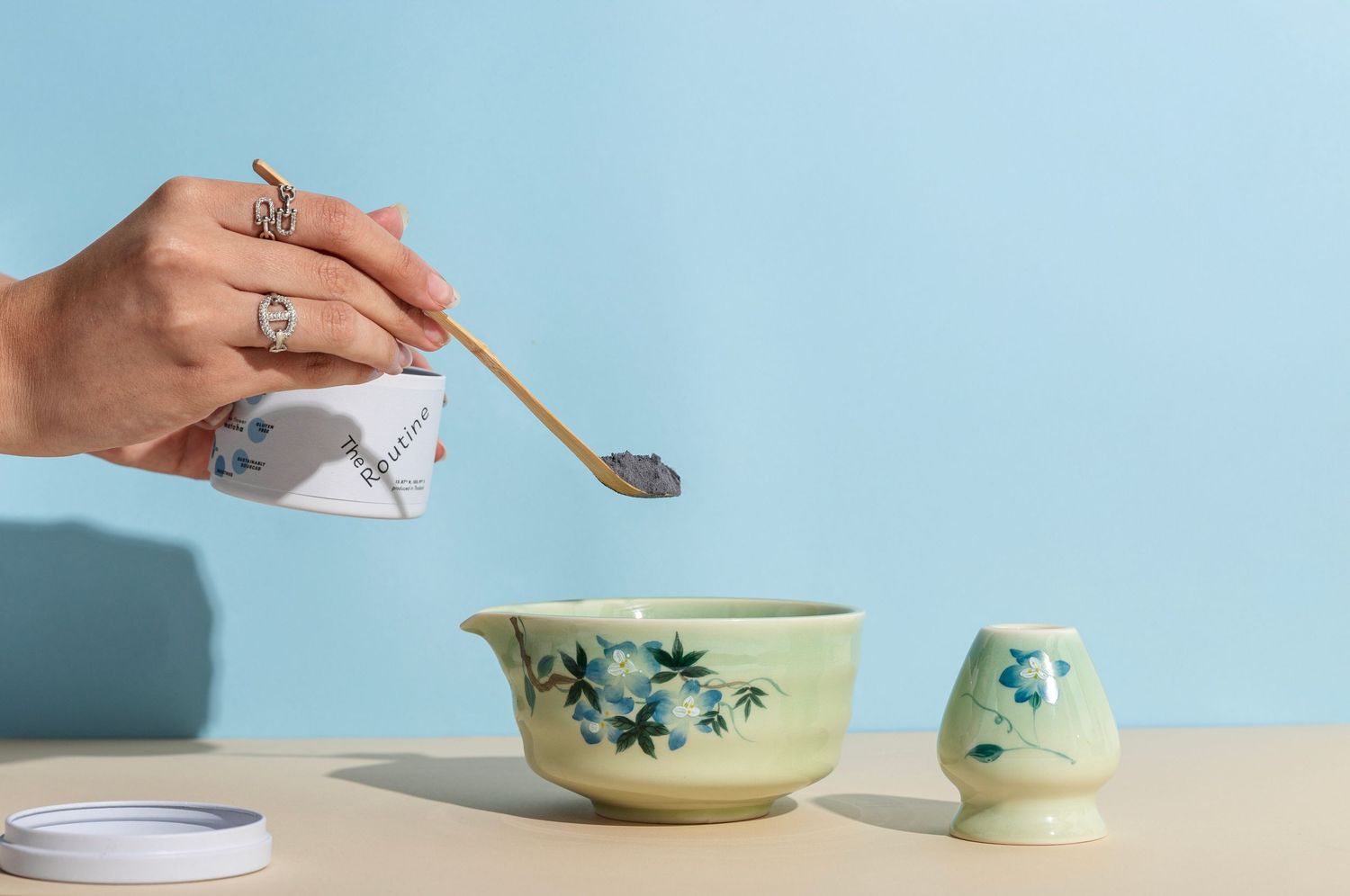 Hands spooning matcha powder into handmade ceramic bowls with matching green glazes on a light surface.