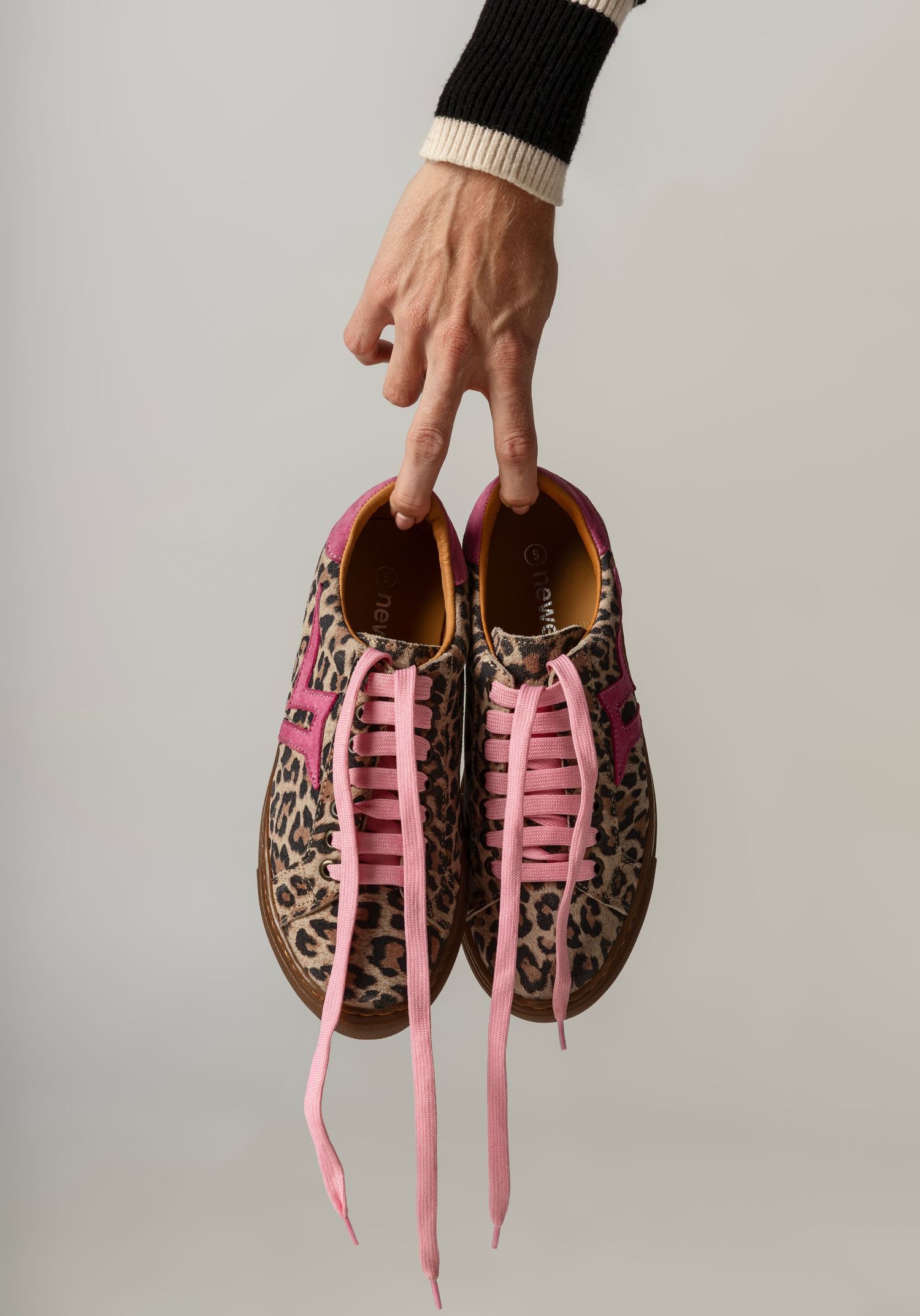 Hand holding leopard print sneakers with pink laces and accents against a neutral background.
